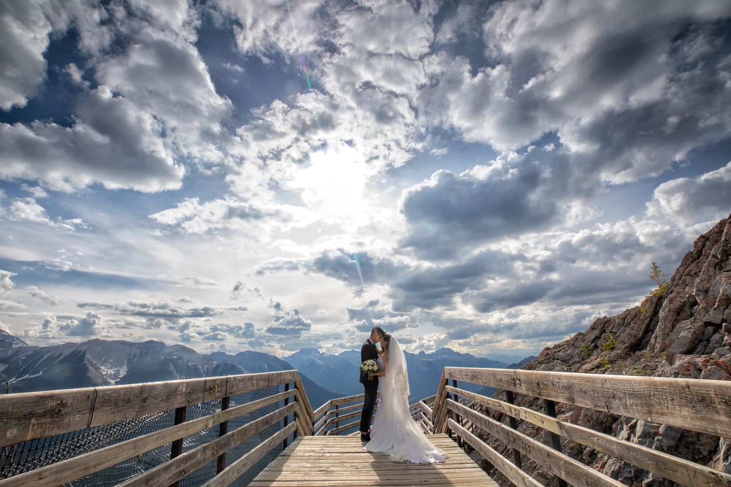 Bride and groom share a romantic kiss on a mountain overlook deck under dramatic cloudy skies.