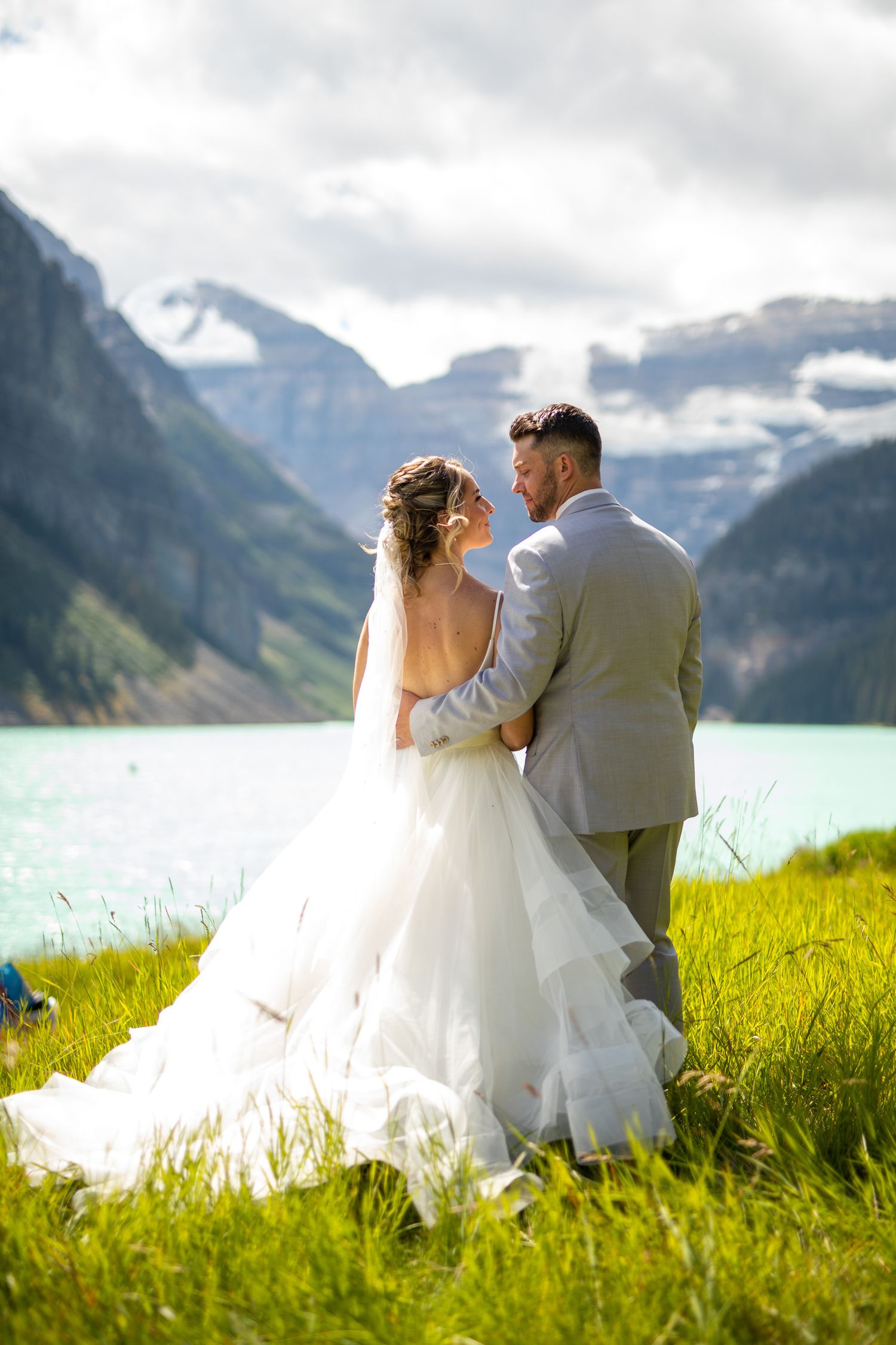 A couple in wedding attire sits in a lush green meadow overlooking the stunning Lake Louise in Banff National Park.