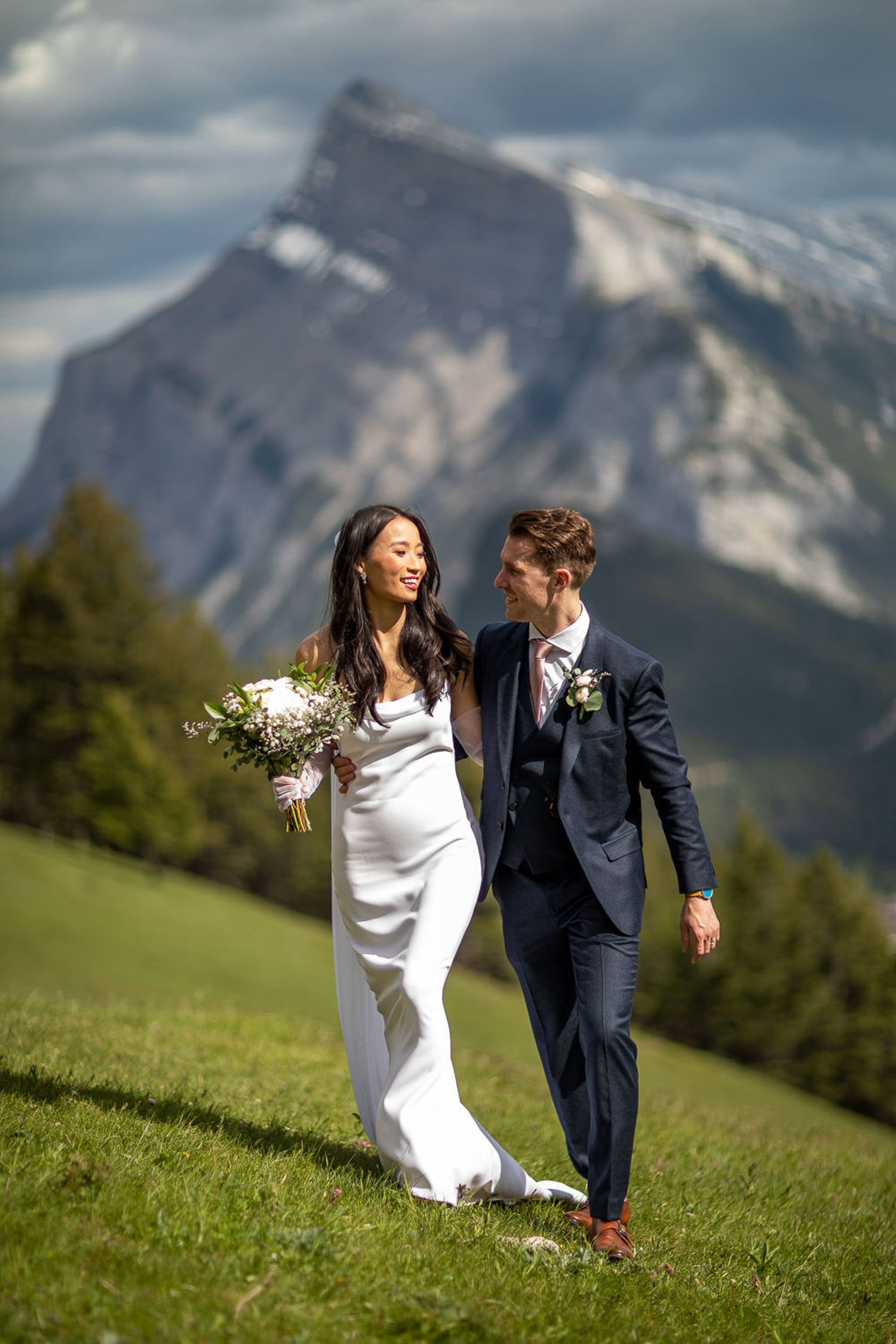 A couple in wedding attire holds hands while walking on a grassy hillside with majestic mountain peaks in the background. Mount NorquayBanff