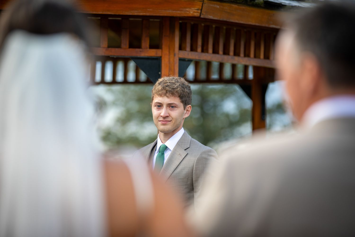 A groom watches his bride during an outdoor wedding ceremony under a wooden pergola. Silvertip resort gazebo in Canmore, Alberta.