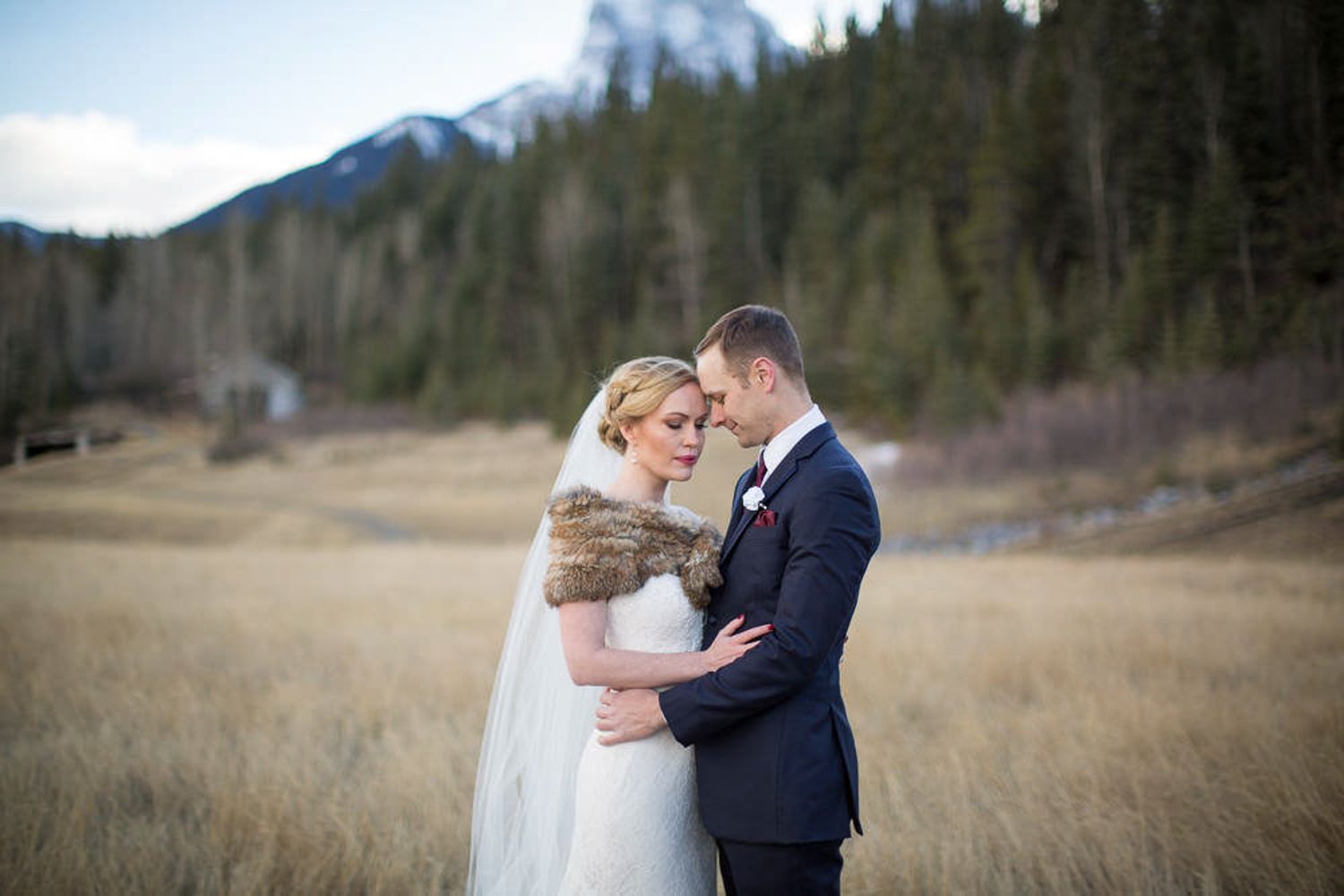 A couple embraces in a scenic mountain landscape with evergreen trees and tall peaks in the background. Canmore mine field.