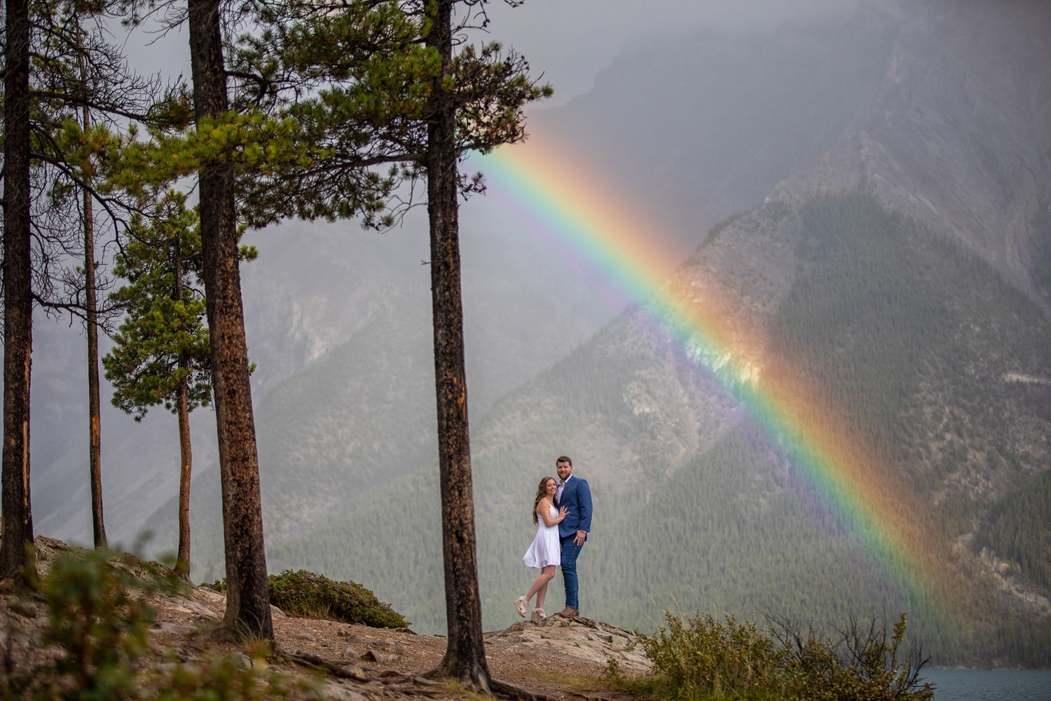 A couple stand on a mountain overlook with a vibrant rainbow arcing across Minnewanka Lake in Banff.