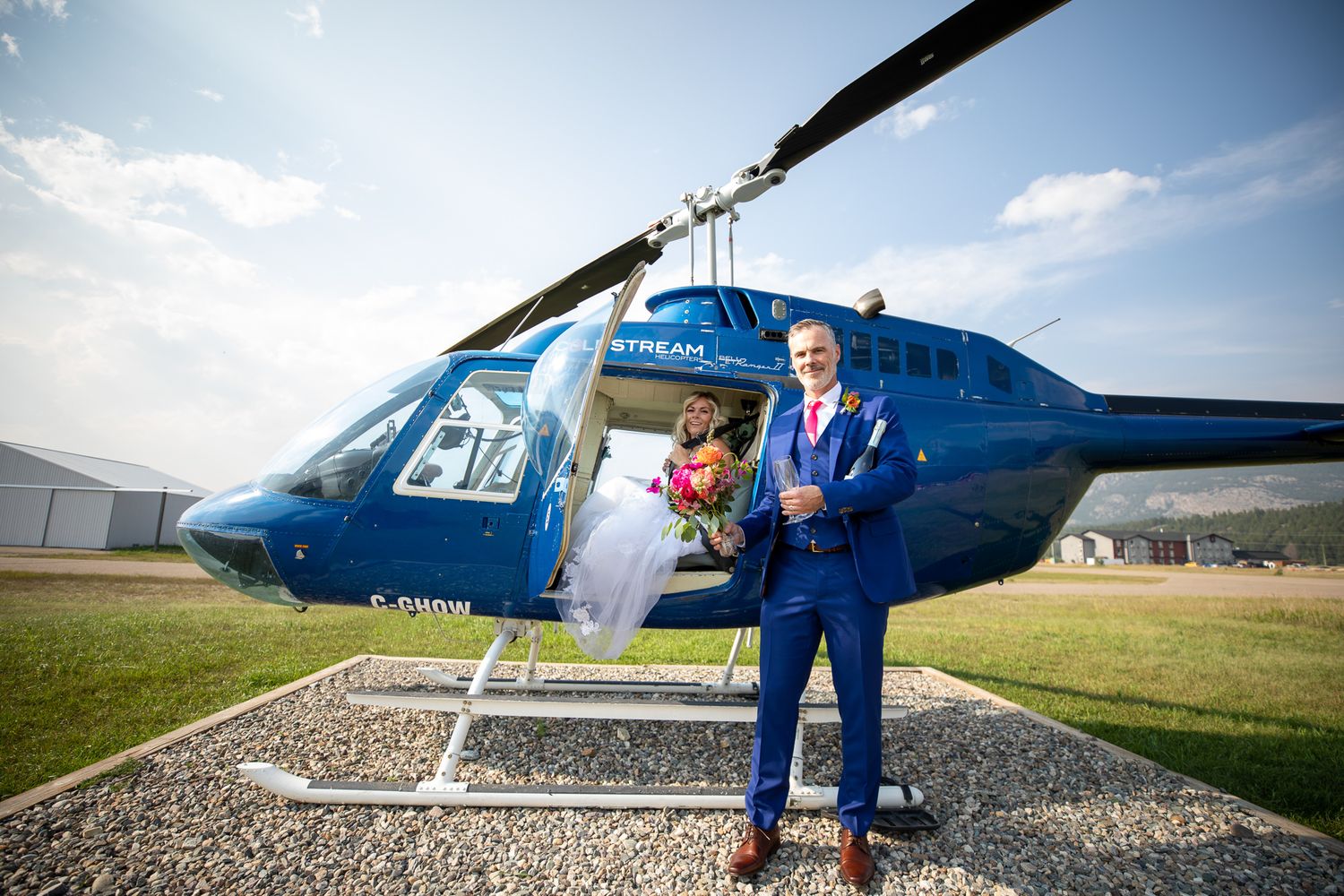 Bride and groom posing beside a blue helicopter on their wedding day at an airfield under a sunny sky Invermere, BC.