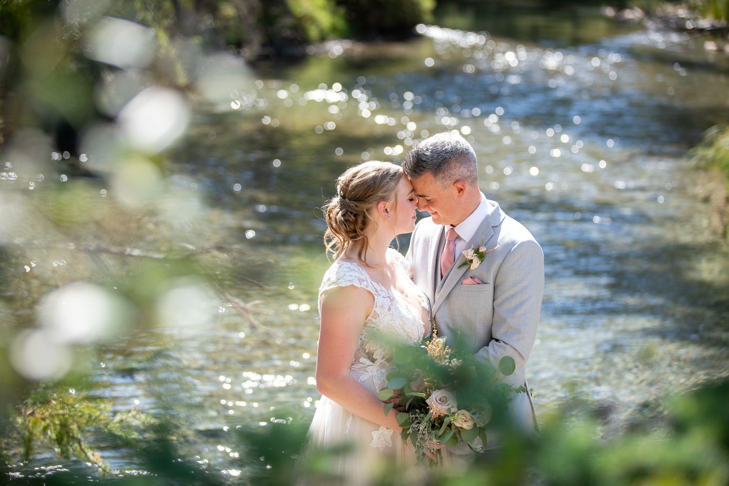 Couple embraces by sparkling stream in romantic outdoor setting with sunlit water and natural greenery in Canmore Ranch, Canmore Alberta