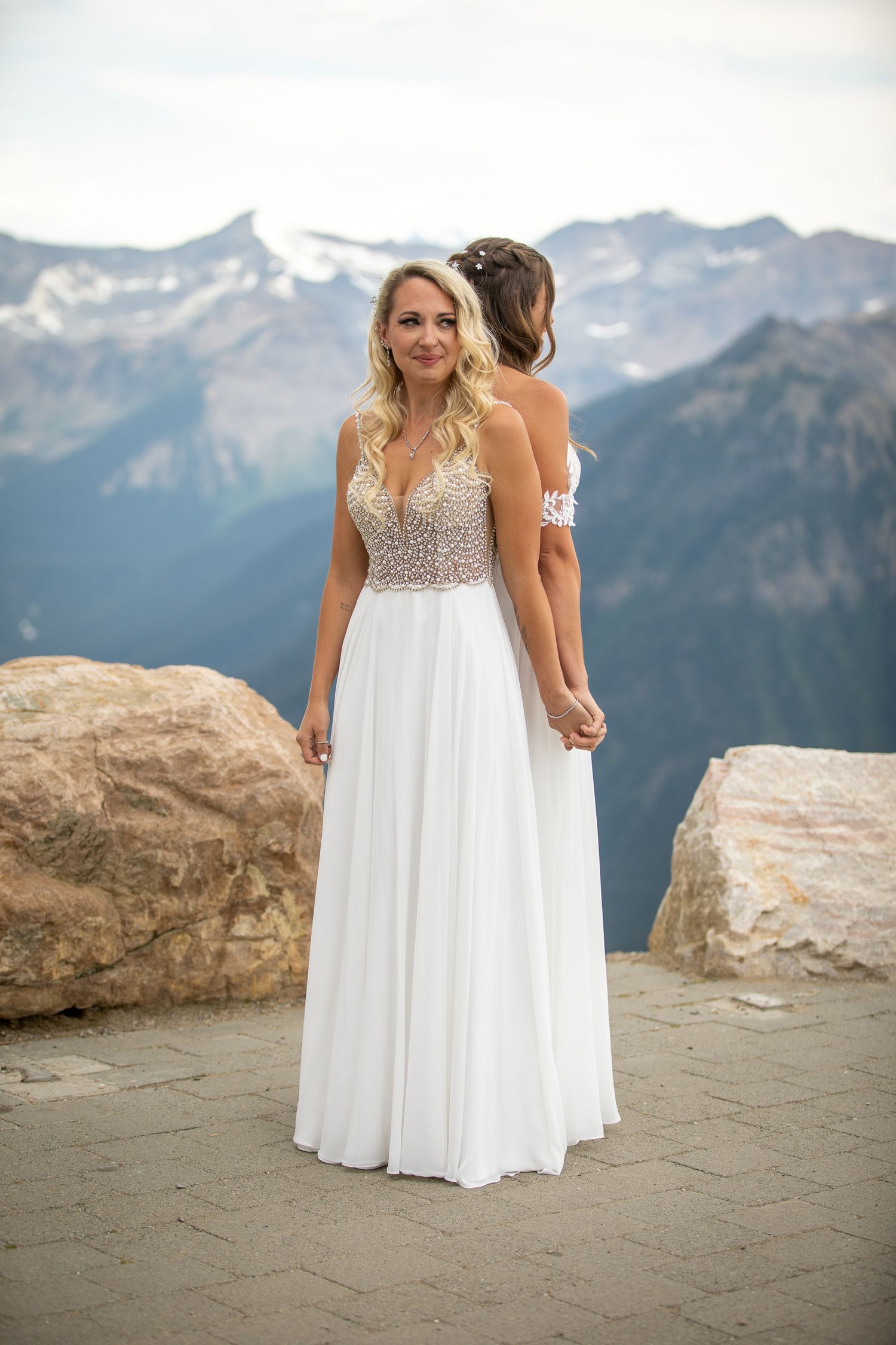 Two brides in white wedding gowns hold hands on a mountain overlook with snow-capped peaks in the background at Kicking Horse Resort, BC
