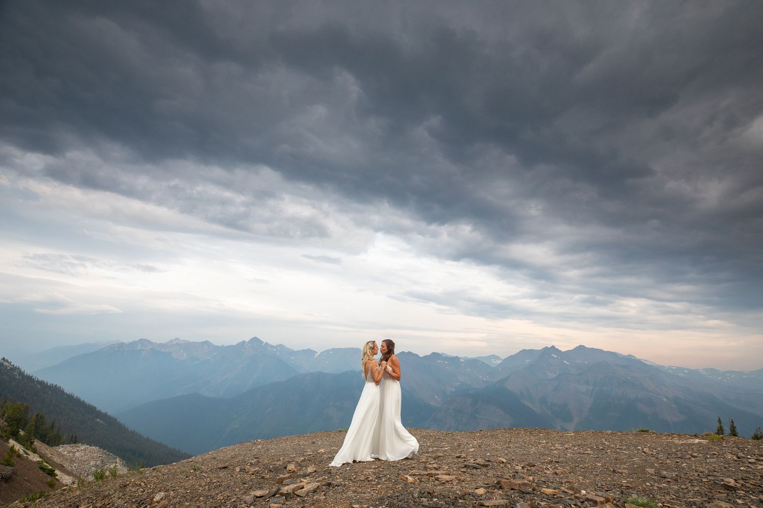 Two brides in wedding attire embraces on a mountain peak under dramatic stormy clouds at sunset at Kicking Horse resort in Golden, BC