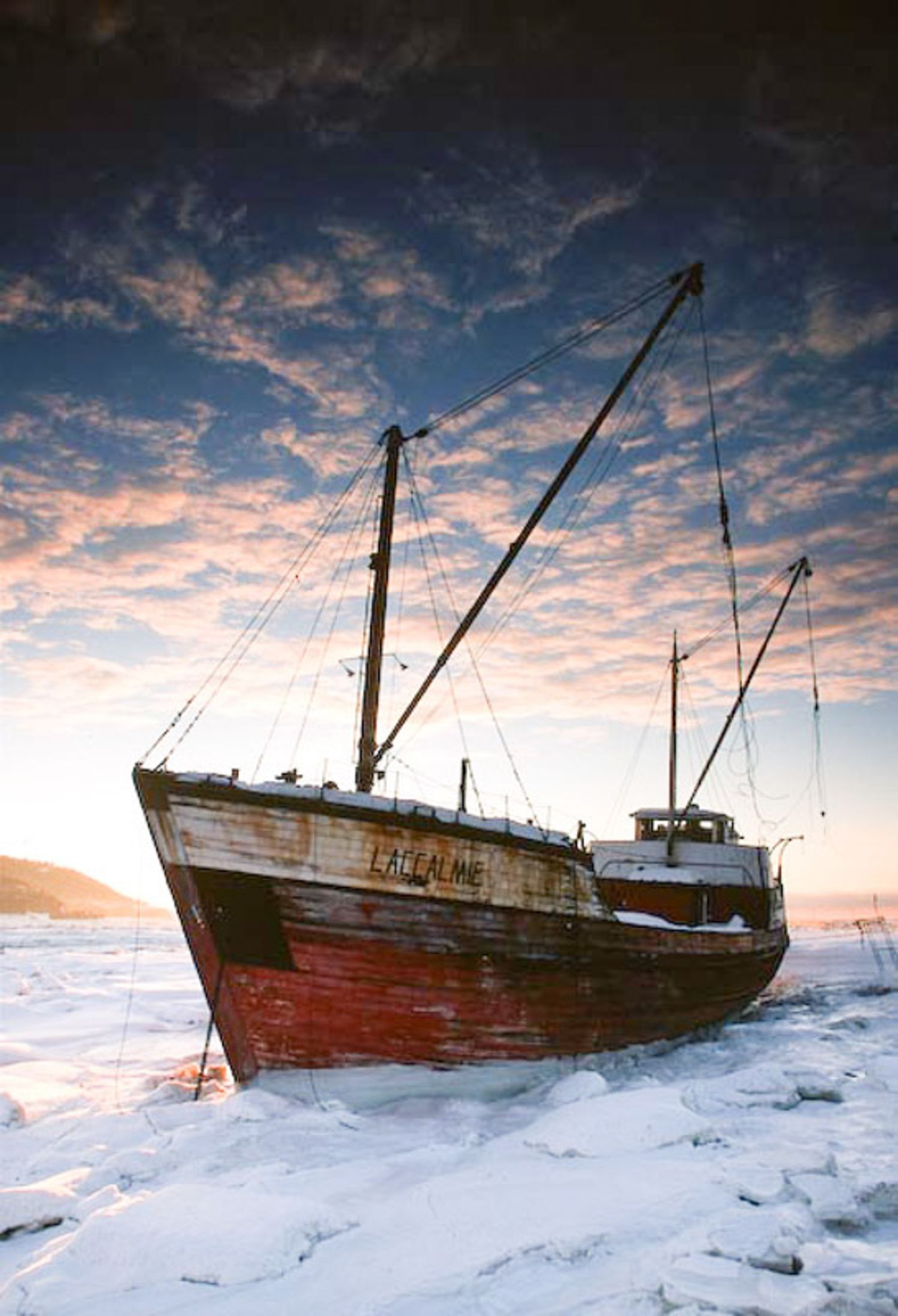 An old wooden fishing boat with red hull stranded in frozen waters against a dramatic sunset sky with pink clouds. Baie-St-Paul, Quebec.