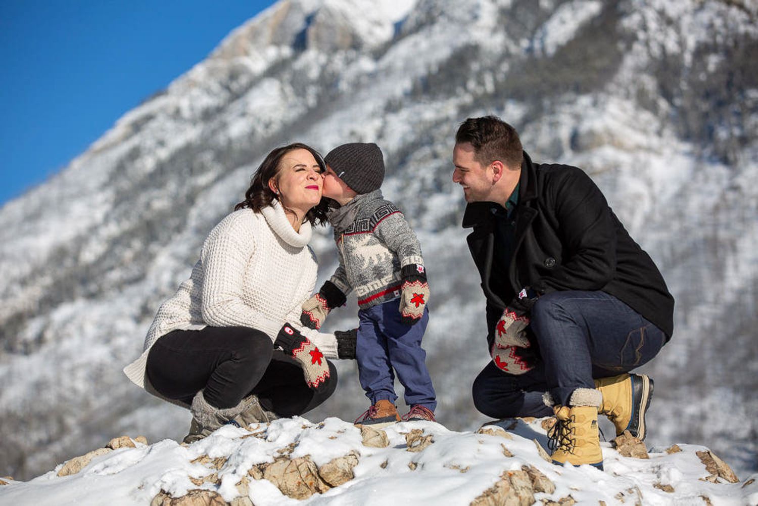 A couple shares a romantic moment on snowy mountain peaks with scenic winter landscape in the background at Minnewanka Lake in Banff, AB.