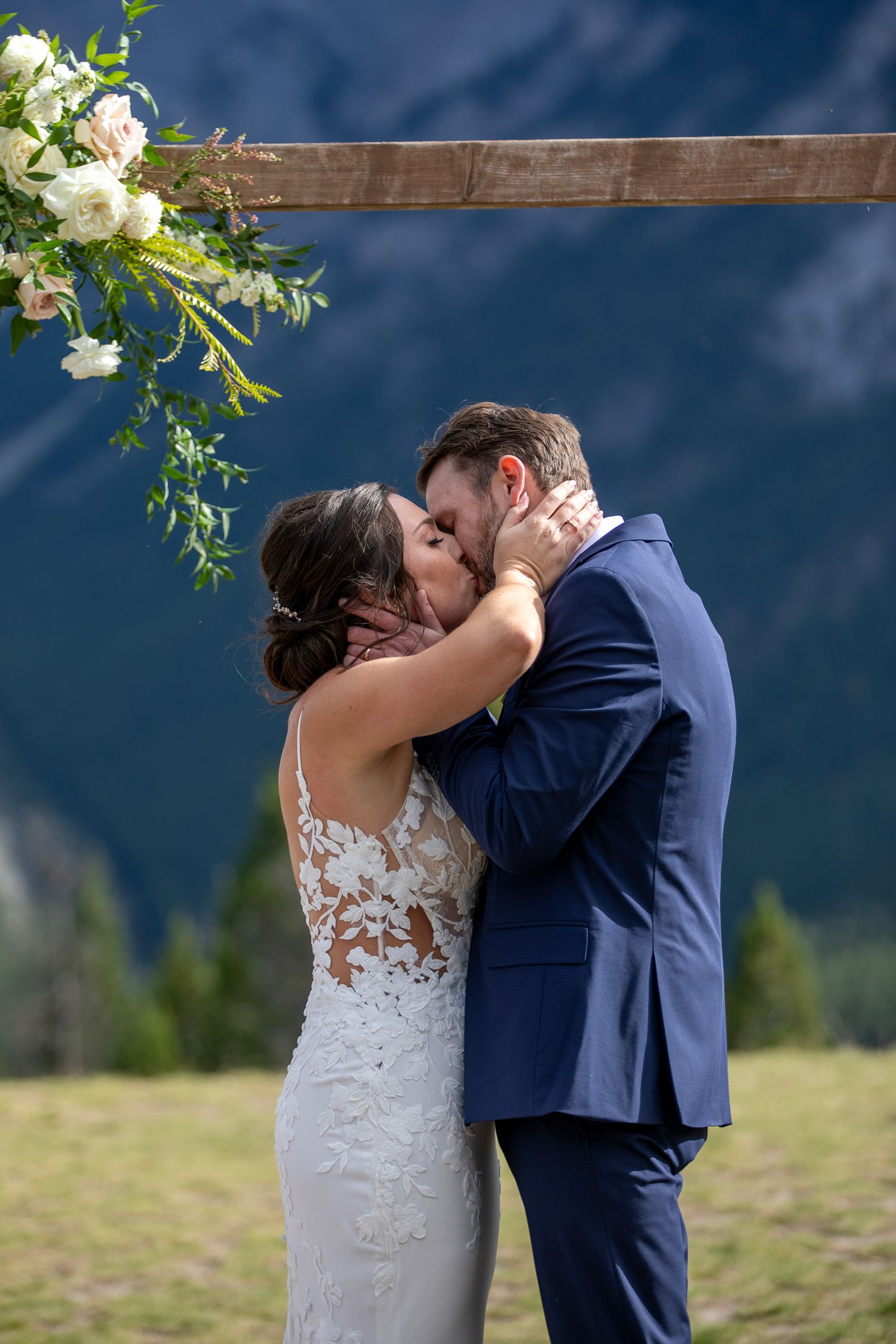 A couple shares a romantic kiss in a mountain meadow during their wedding ceremony with white floral decorations. Banff, Alberta