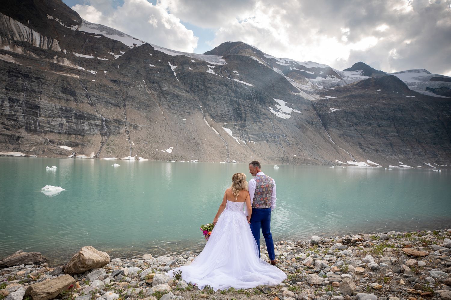 Bride and groom stand at turquoise glacial lake shore with dramatic snow-capped mountain backdrop on their wedding day. Panorama, BC.