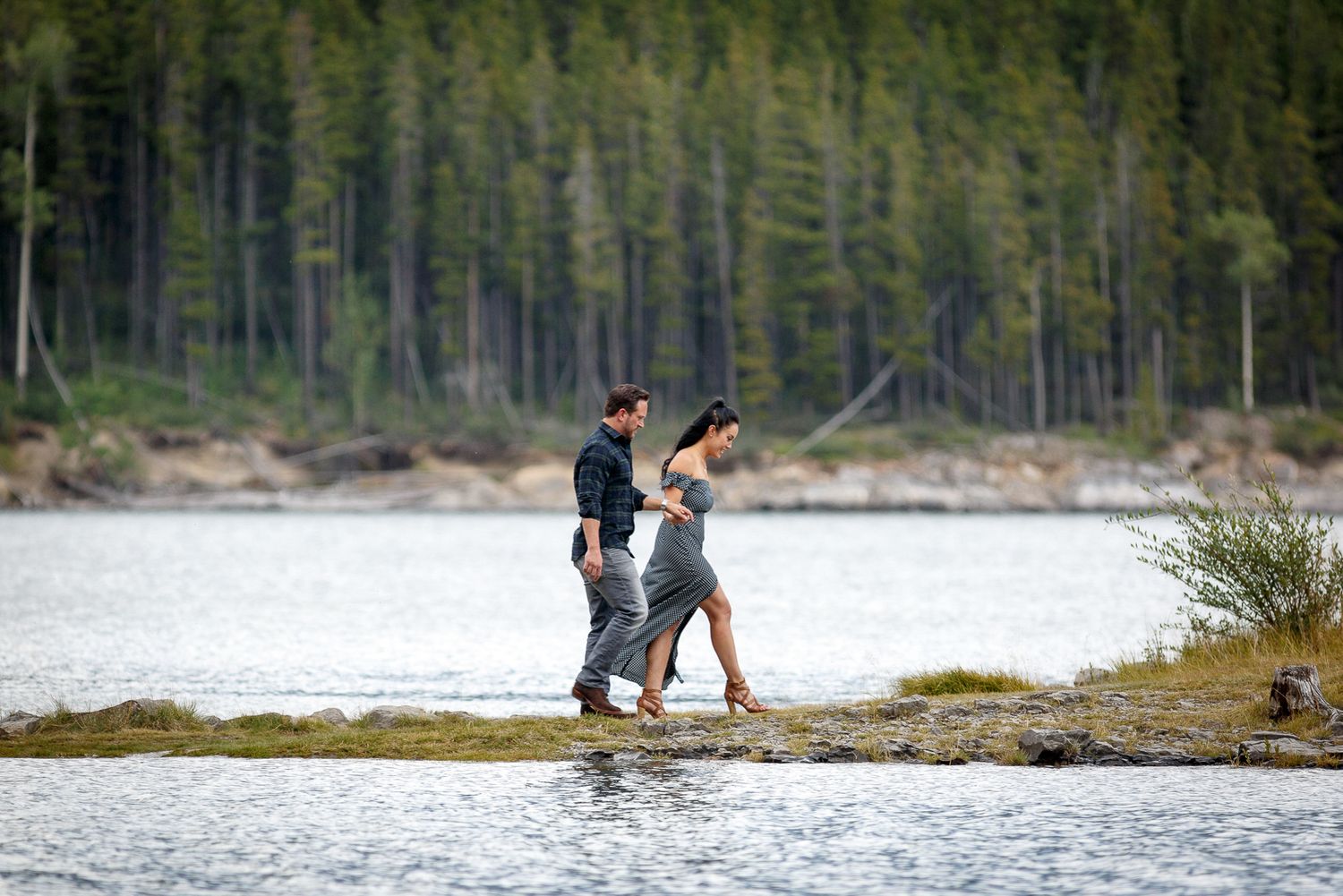 Two people walking together in shallow water near a forested shoreline at Lake Minnewanka in Banff.