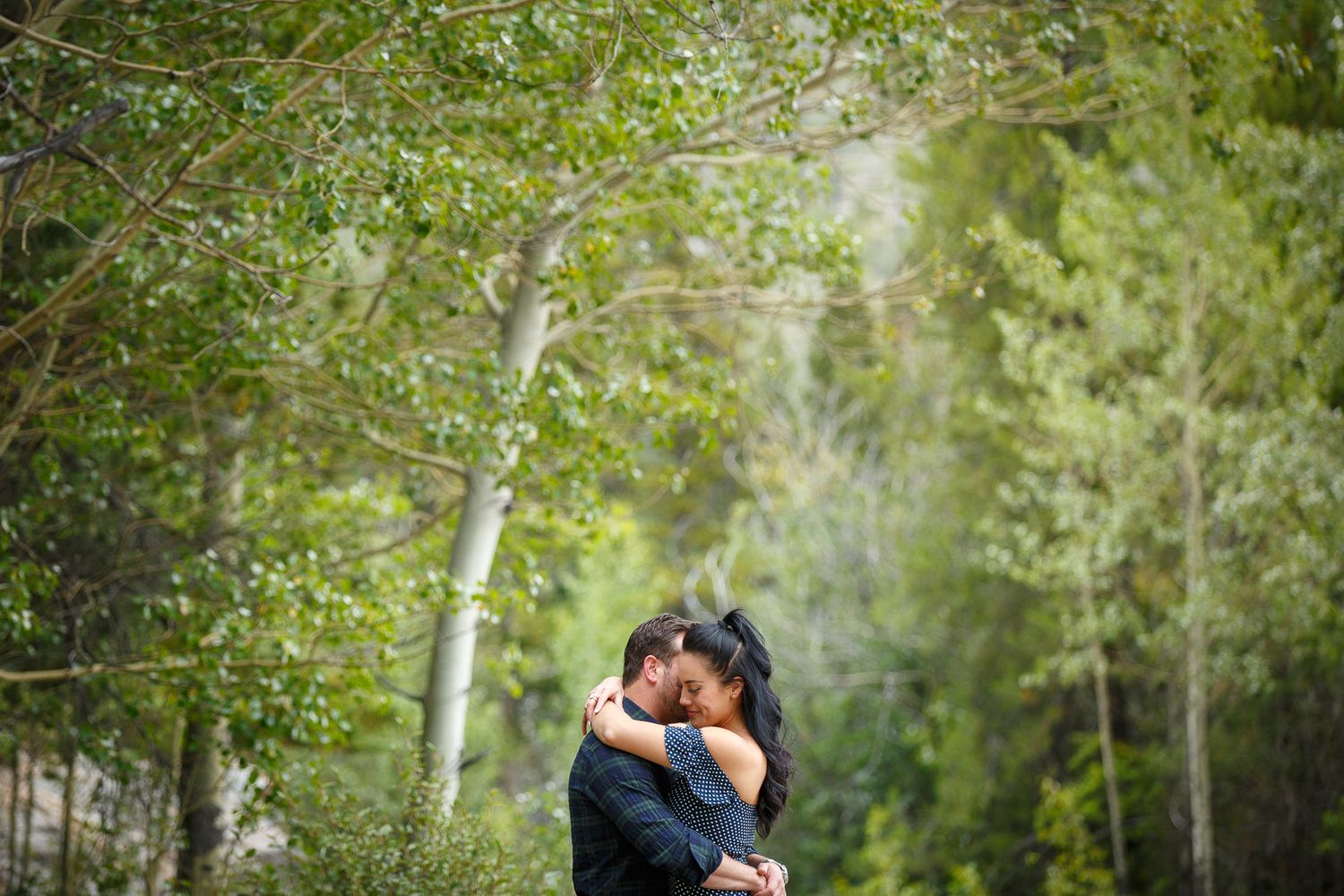 A couple embracing in a lush, green forest setting at Lake Minnewanka in Banff.