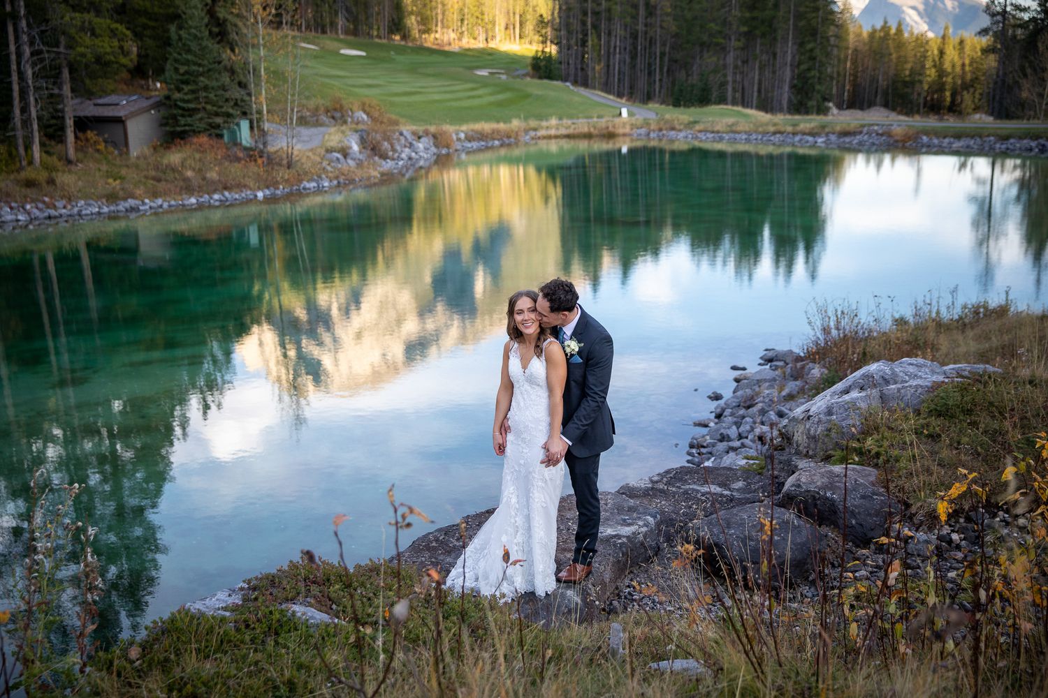 A couple poses near a serene mountain lake surrounded by evergreen trees during golden hour at sunset at Silvertip resort in Canmore, AB