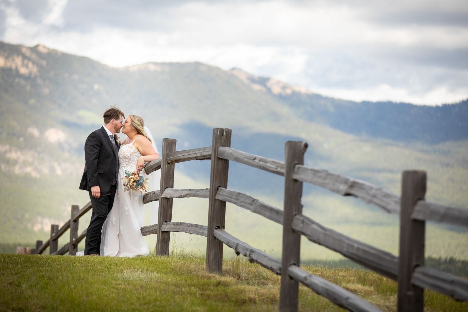 A couple shares a romantic moment along a rustic wooden fence with rolling mountains in the background. Brady Creek ranch. Invermere, BC.