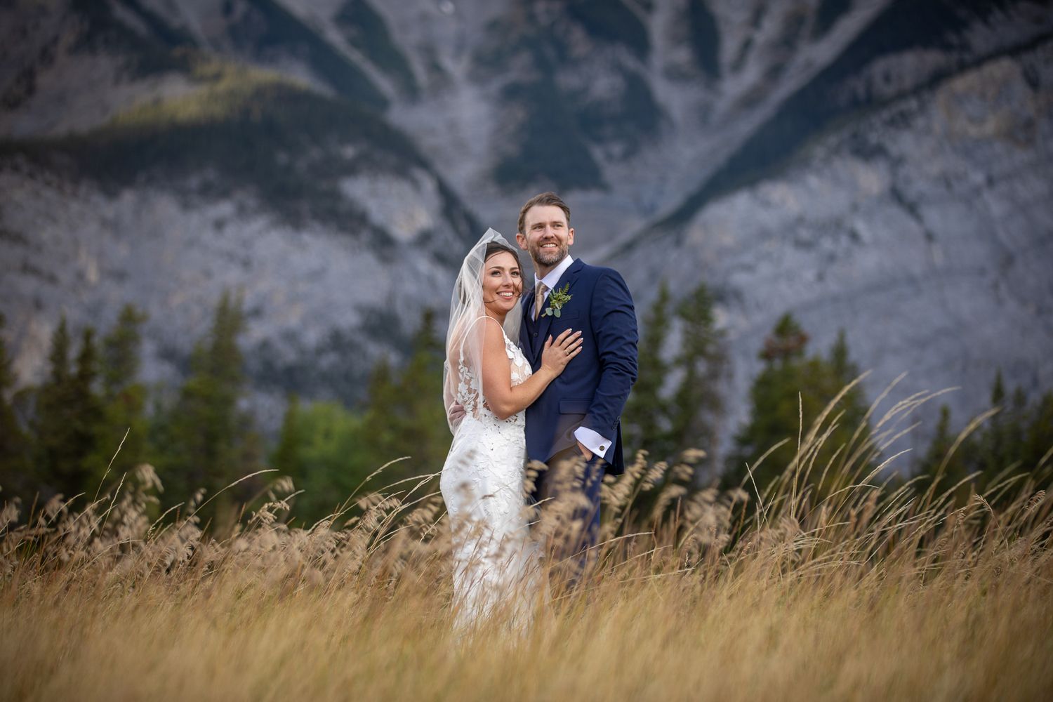 A couple in wedding attire embraces in a field with majestic mountain peaks in the background at sunset in Banff. Banff National Park.