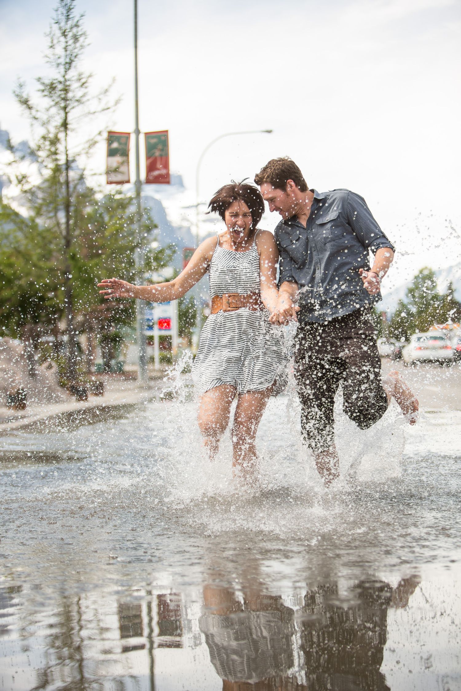A couple splashes joyfully through a puddle on a rainy day, their reflection visible in the water below. Canmore Alberta