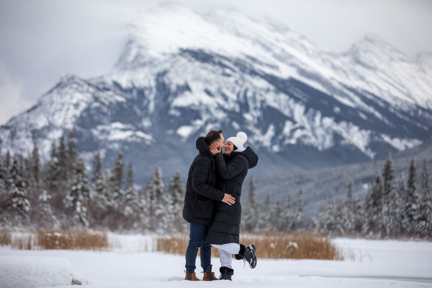 A couple embraces in a romantic winter engagement photoshoot with snow-capped mount Rundle in the background in Banff, AB.