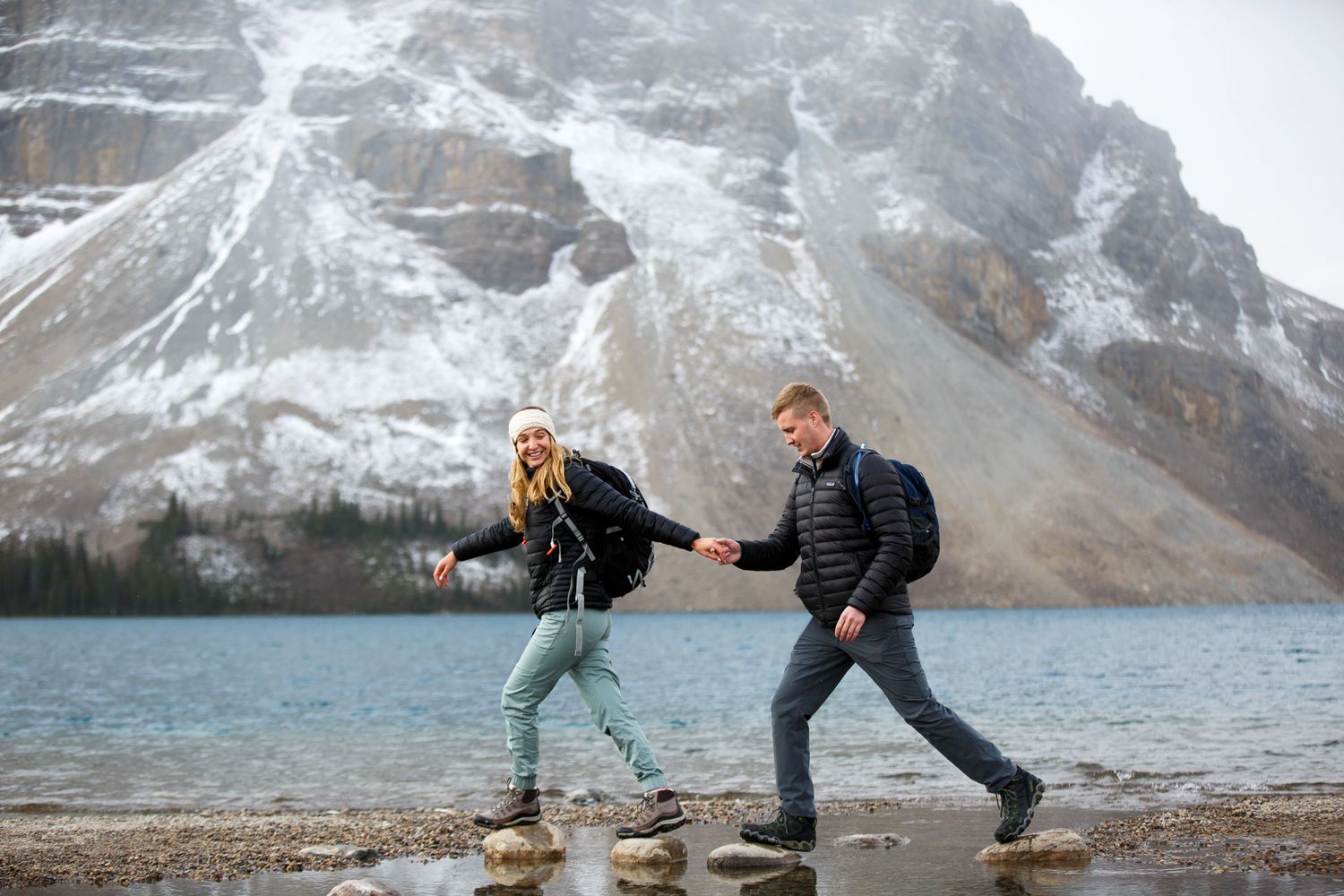 Two people playfully dance and pose in front of snow-covered mountains at Moraine Lake in Banff National Park.