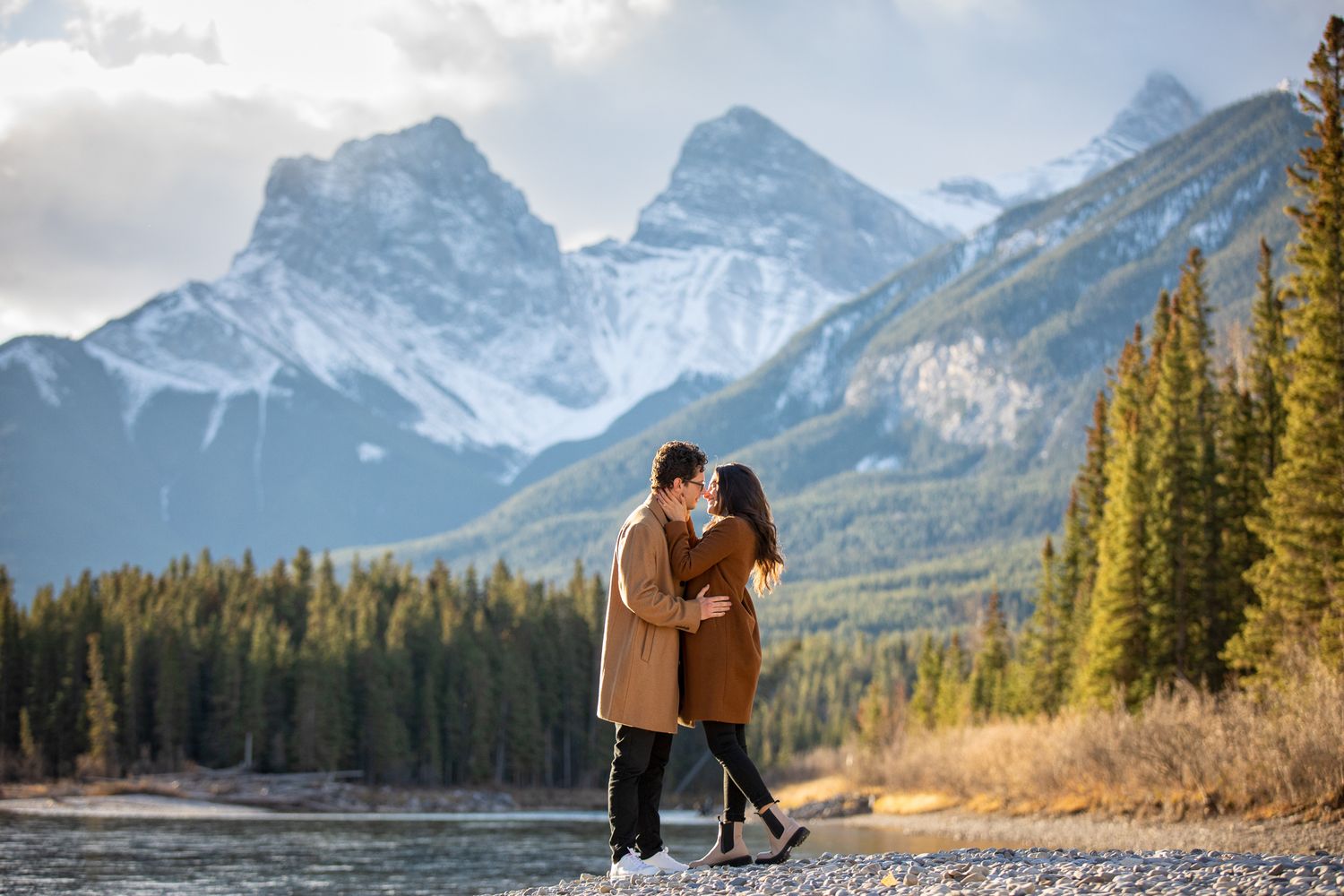 A couple shares an intimate moment against a backdrop of snow-capped Three Sisters mountains and autumn trees in Canmore, AB.