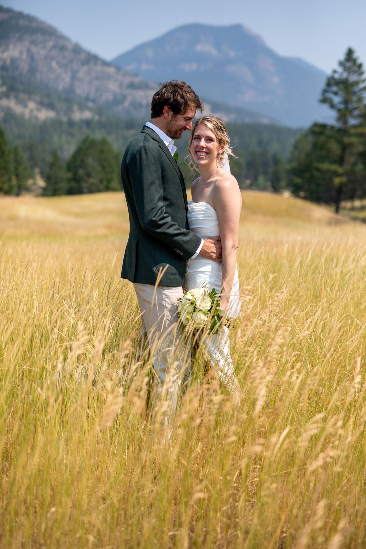 Bride and groom embracing in golden meadow with mountain backdrop on sunny wedding day at Swansea Ranch in Invermere, BC