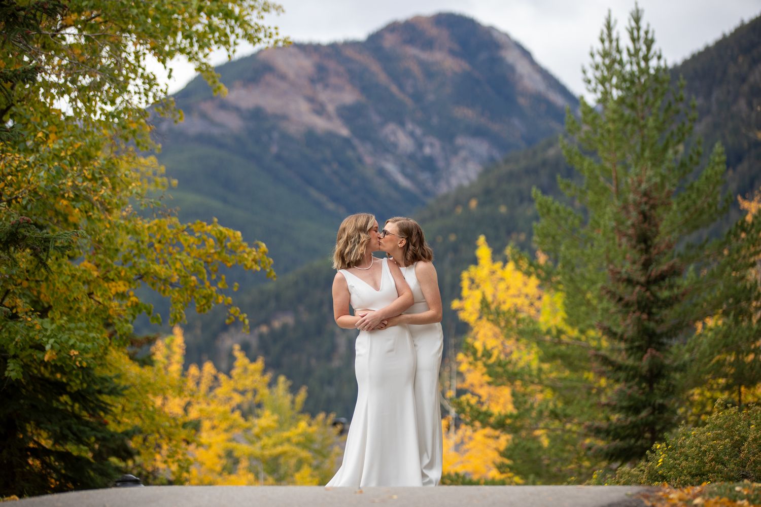 A couple shares a romantic moment against a backdrop of mountains and golden autumn foliage in a scenic outdoor setting. Panorama BC