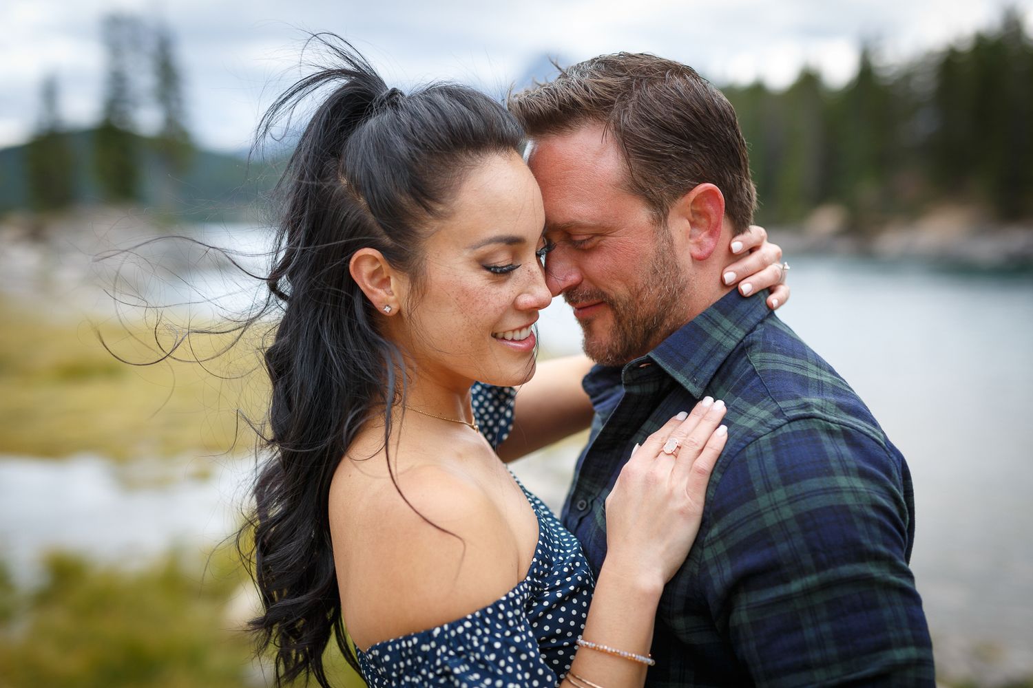 Couple sharing a romantic moment by a mountain lake, woman in floral dress with flowing dark hair, man in plaid shirt at Lake Minnewanka.