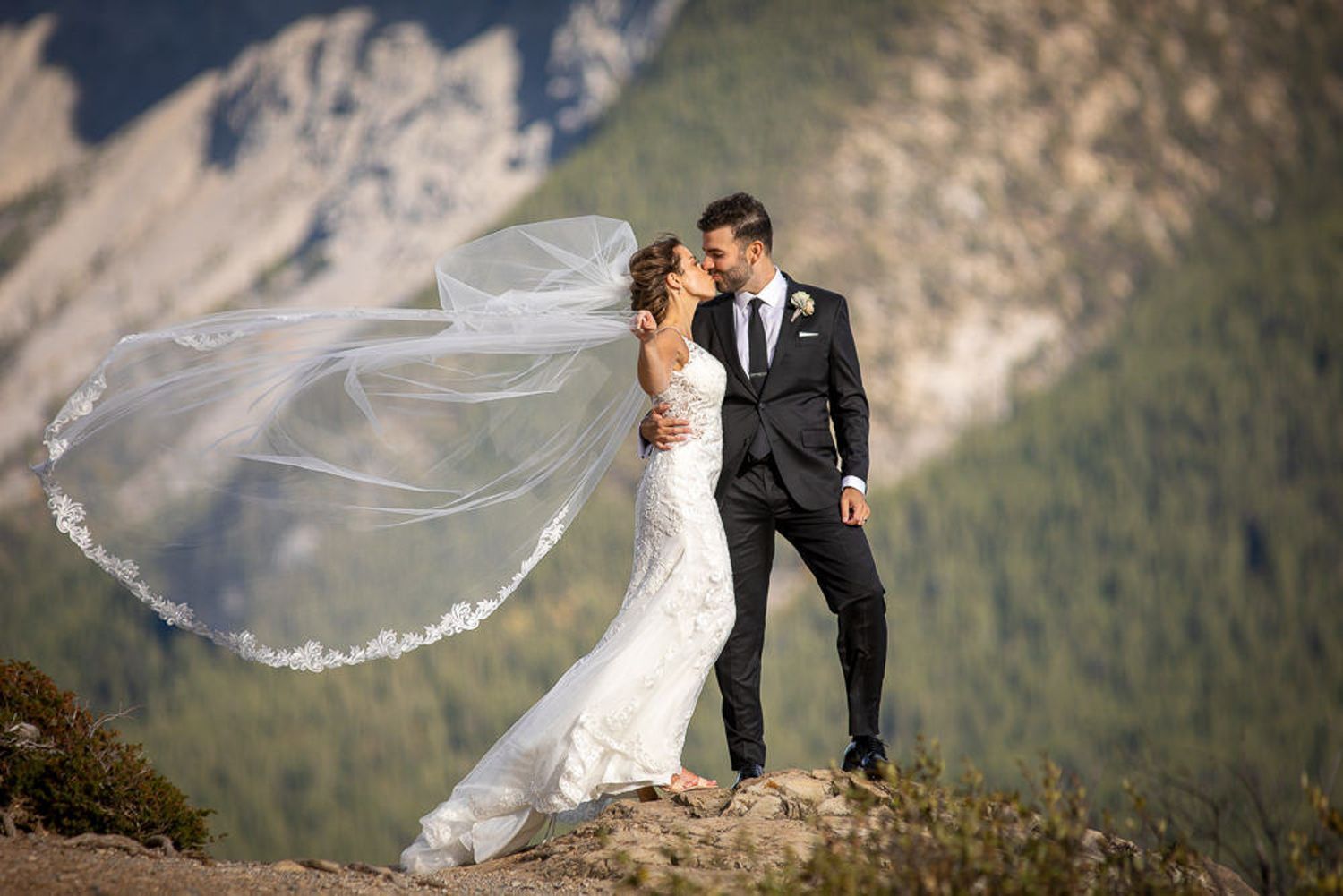 A flowing white wedding veil catches the mountain breeze as figures stand against a dramatic alpine backdrop. Banff Minnewanka Lake.