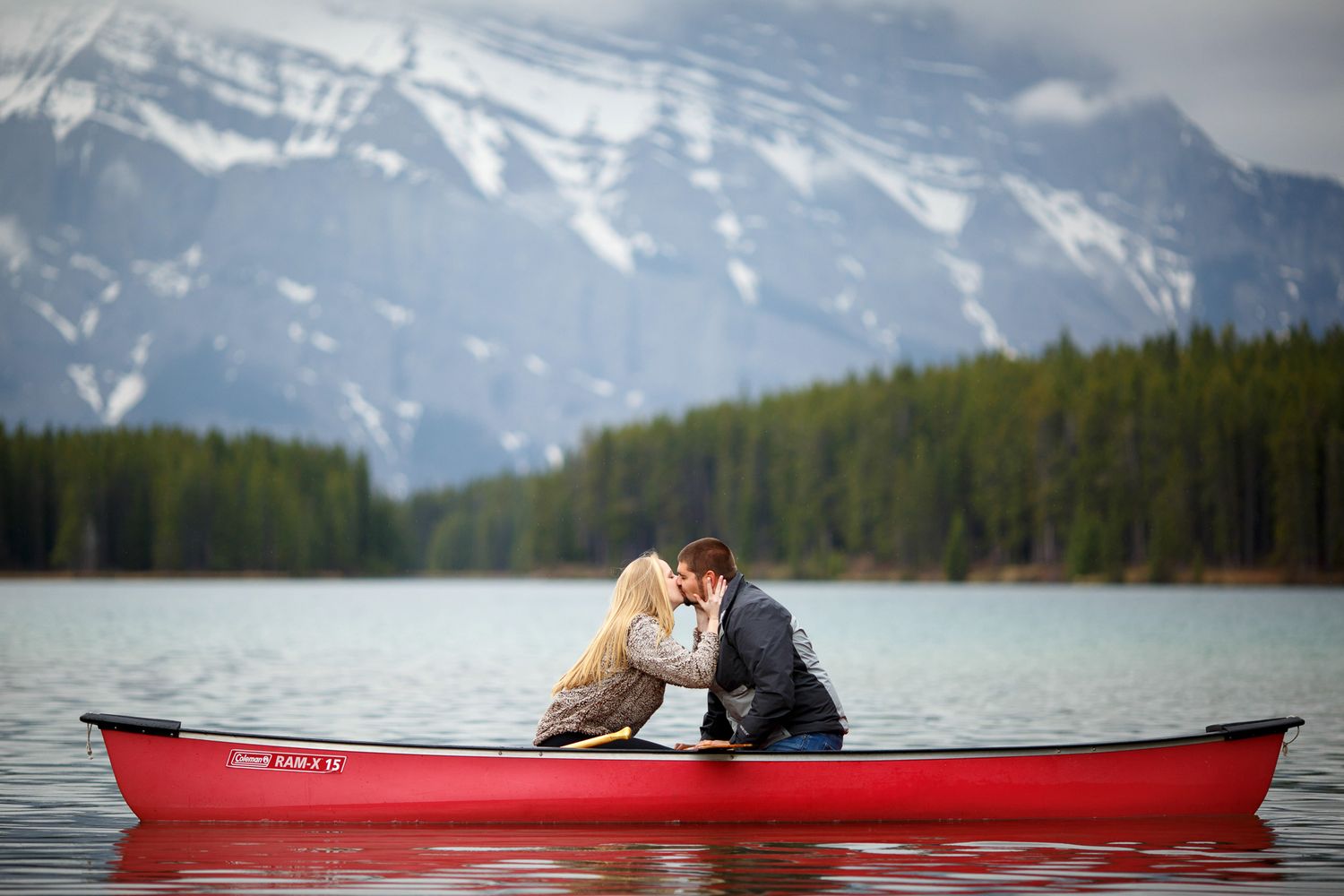 A couple shares a romantic moment in a red canoe on a scenic Two Jack lake surrounded by evergreen forests and Mount Rundle in background.