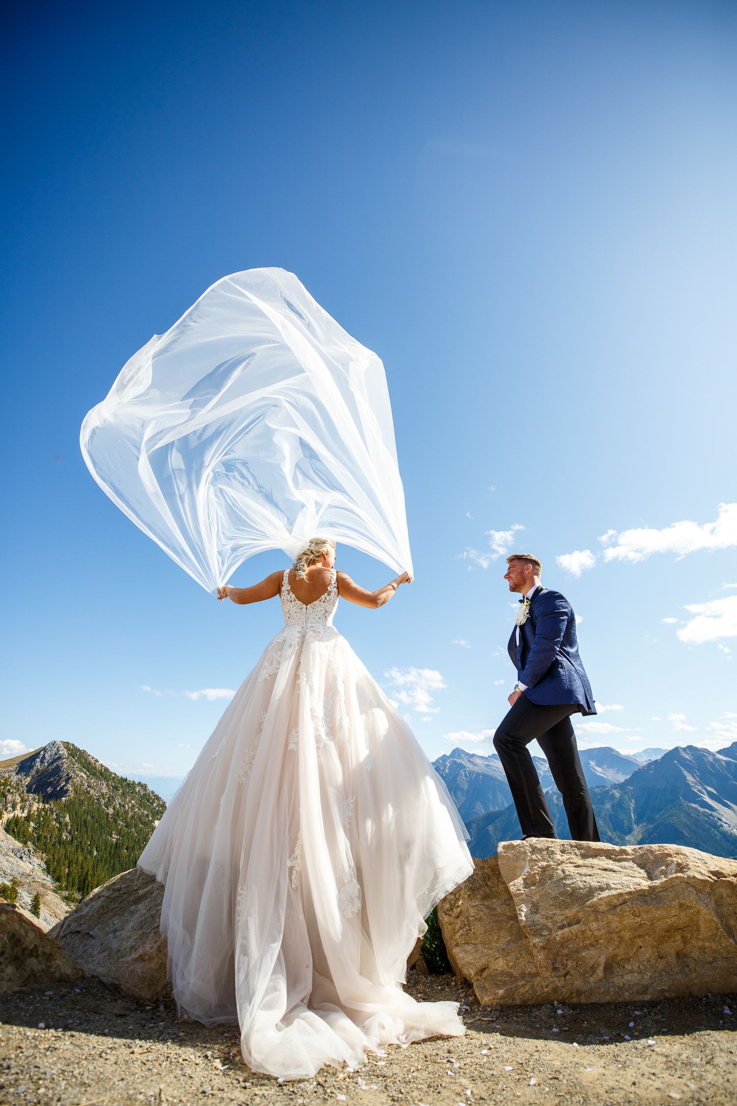 A wedding couple poses dramatically on mountain rocks with flowing white dress and veil against blue sky and mountain peaks.Golden, BC.