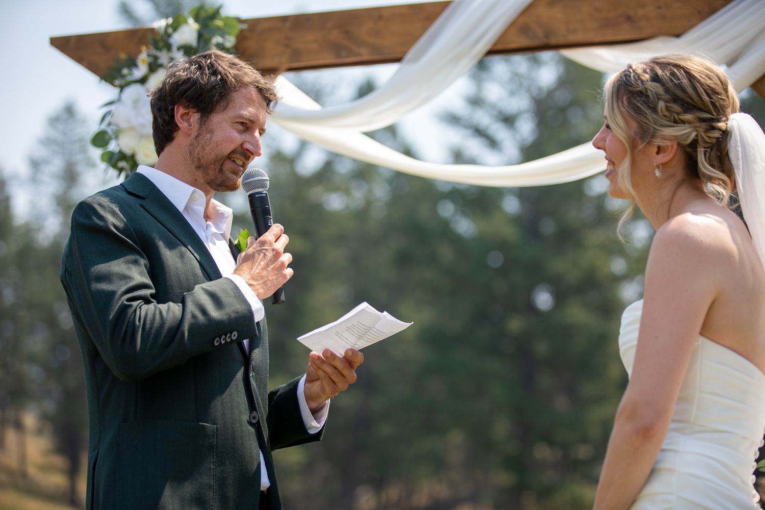Groom reads wedding vows from paper while holding microphone outdoors under wooden arch with white draping at Swansea Ranch in Invermere, BC