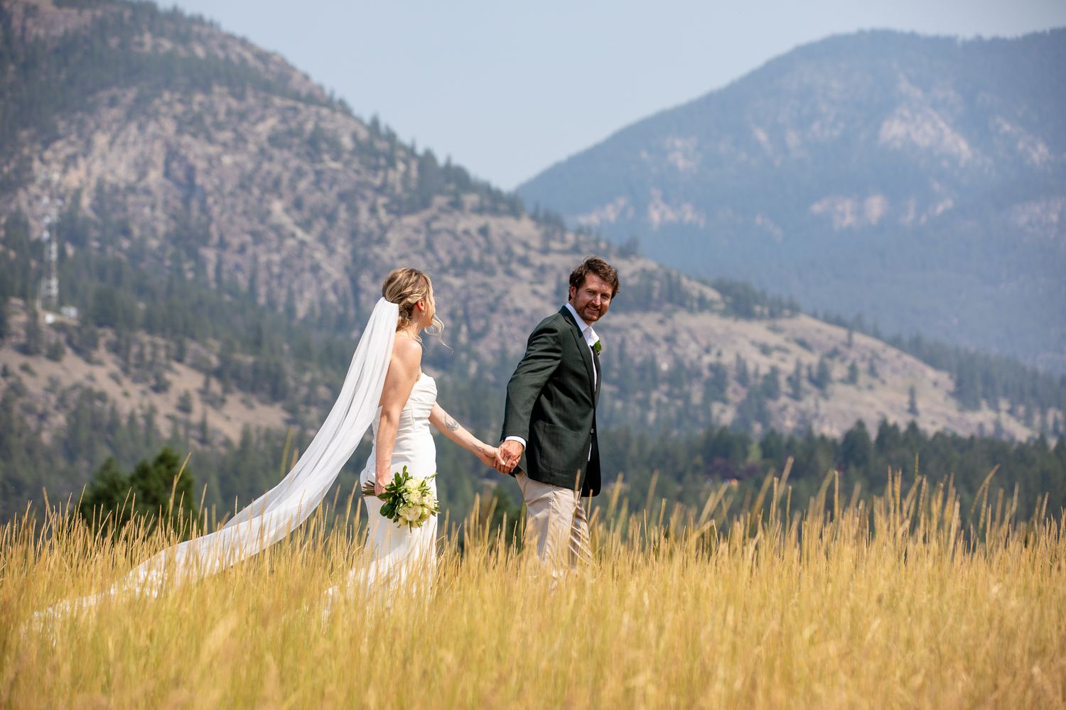 Bride and groom holding hands in golden meadow with mountain forest backdrop on their wedding day at Swansea Ranch in Invermere, BC