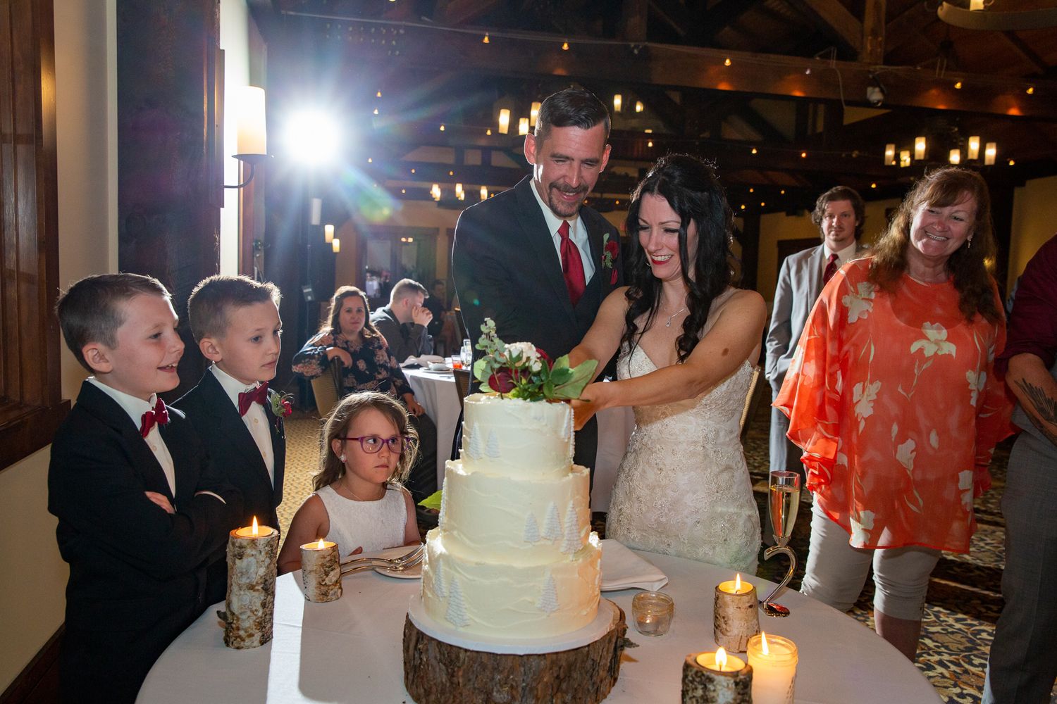 Bride and groom cutting white wedding cake on wood stand, surrounded by children and guests in elegant reception at Silvertip resort.