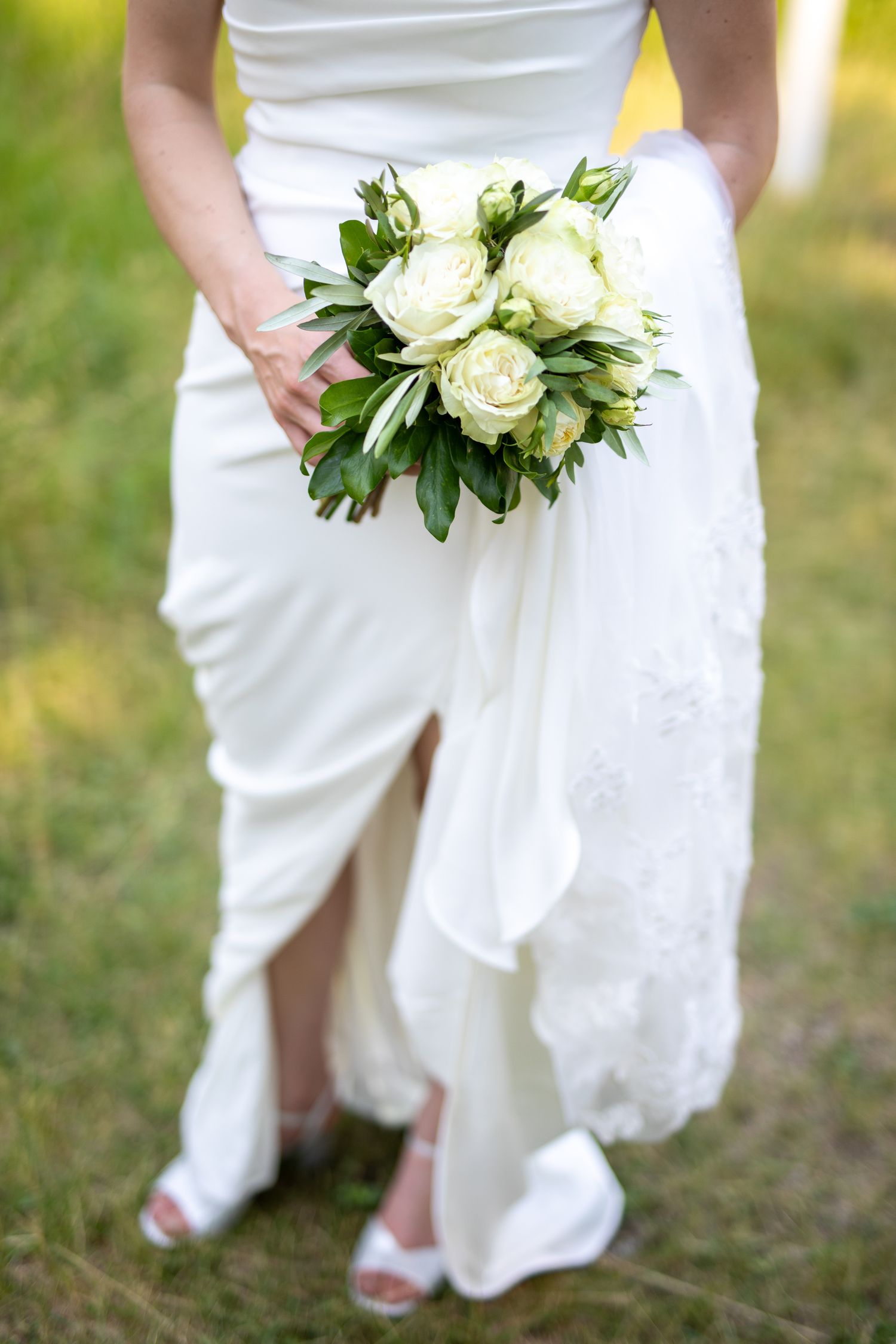 Bride in white wedding dress holding elegant bouquet of white roses and greenery outdoors on green grass at Swansea Ranch in Invermere, BC
