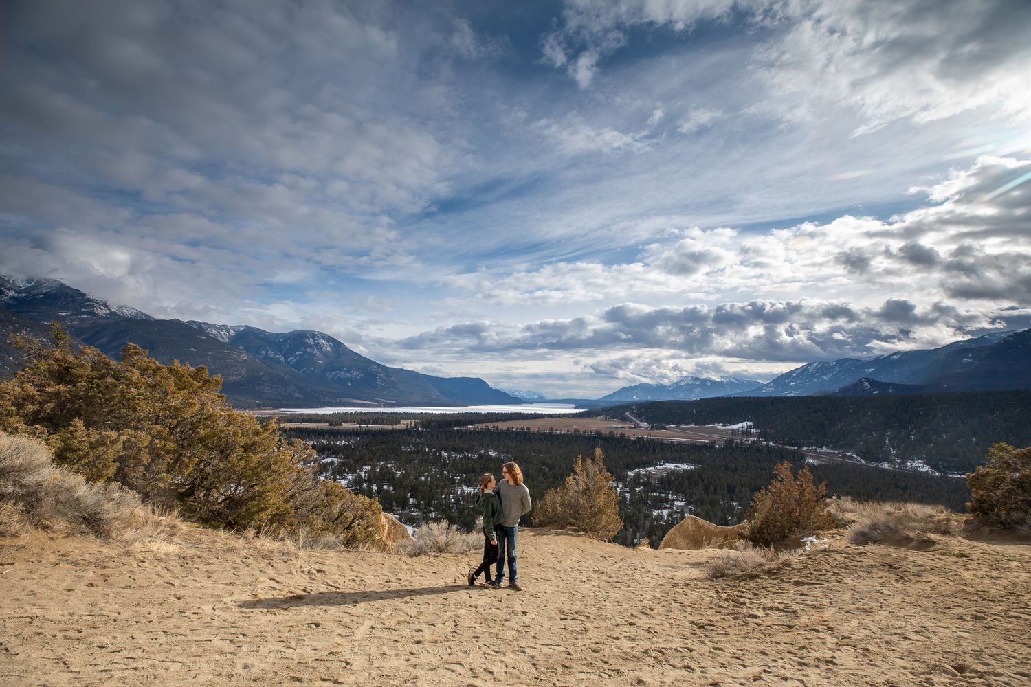 Newly engaged couple stand on sandy trail overlooking Columbia lake with dramatic cloudy sky at Fairmont Hot Springs, BC
