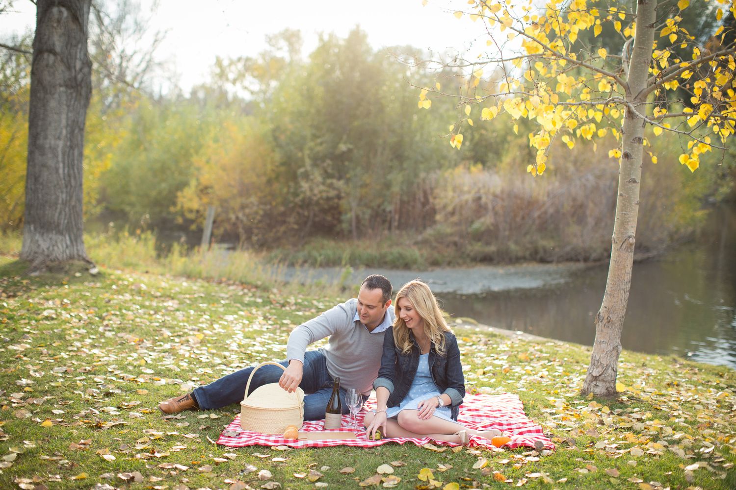 A couple enjoys a picnic on a blanket surrounded by fall foliage near a tranquil pond at sunset in Fish Park, Calgary, AB.