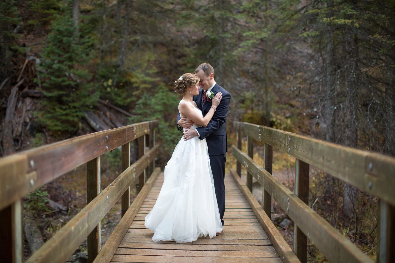 A couple shares a romantic moment on a wooden bridge surrounded by lush evergreen forest. Emerald Lake Lodge, Field BC