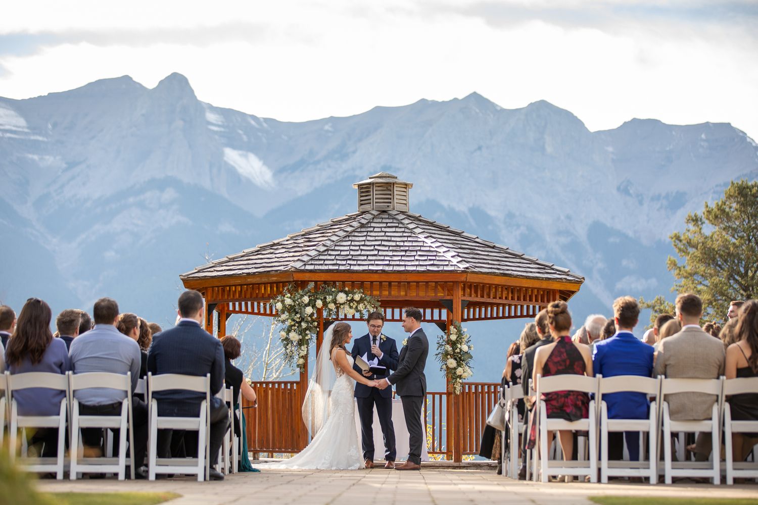 Wedding guests seated in white chairs at an outdoor mountain ceremony with the Silvertip Resort gazebo overlooking scenic peaks Canmore, AB.