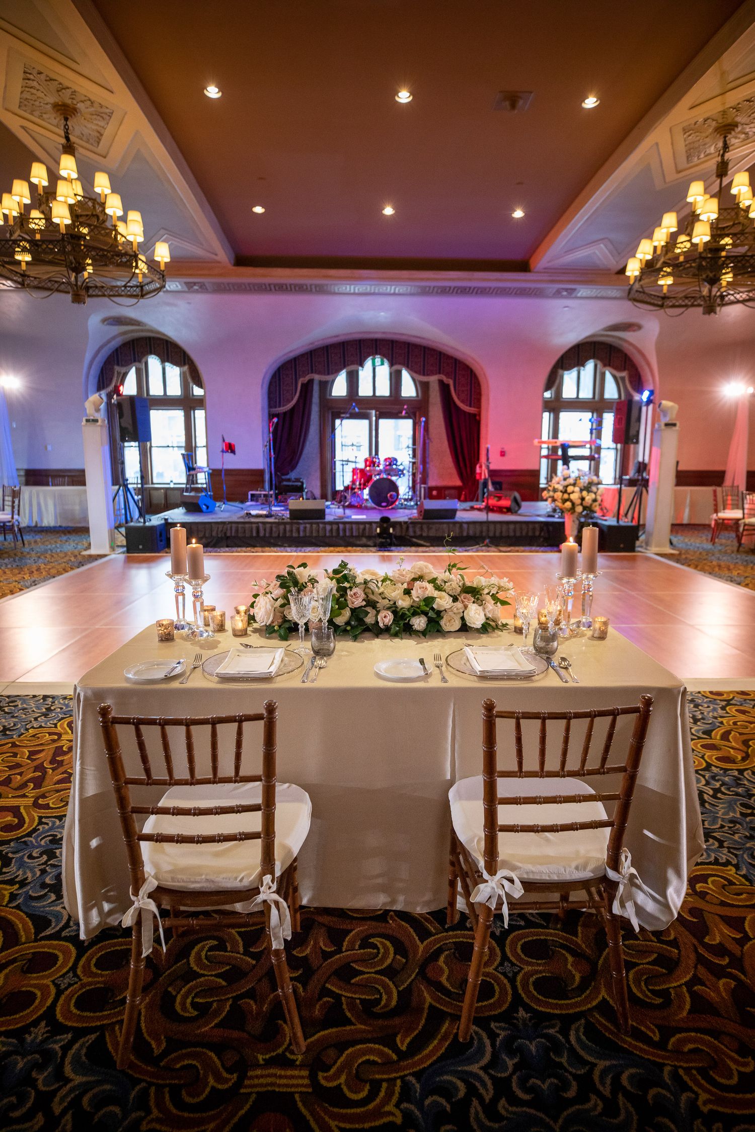 Elegant wedding reception table with white chairs and floral centerpiece in grand ballroom with arched windows and chandeliers.