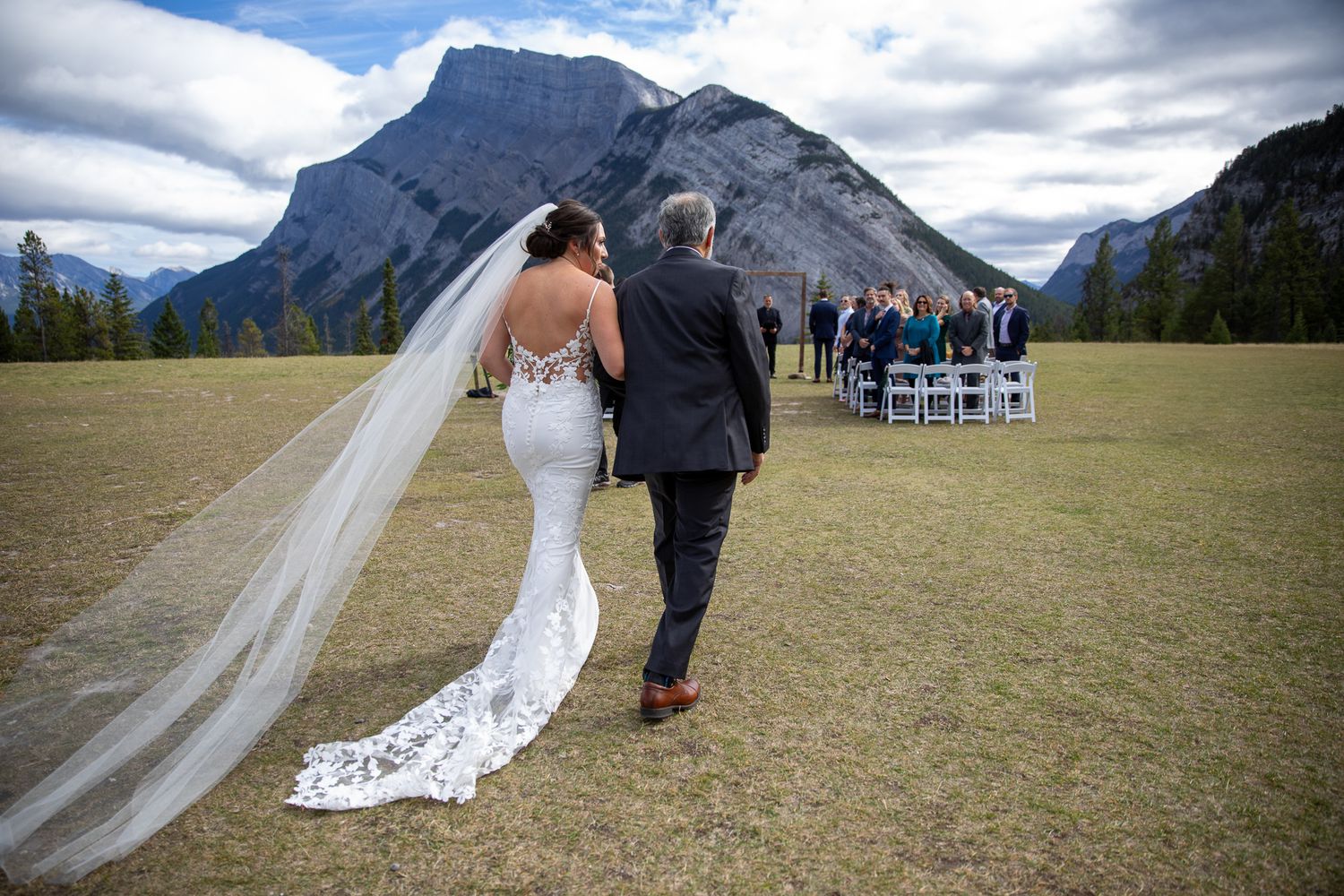 A couple walks towards an outdoor wedding ceremony setup with Mount Rundle rising majestically in Banff National Park. Banff, AB