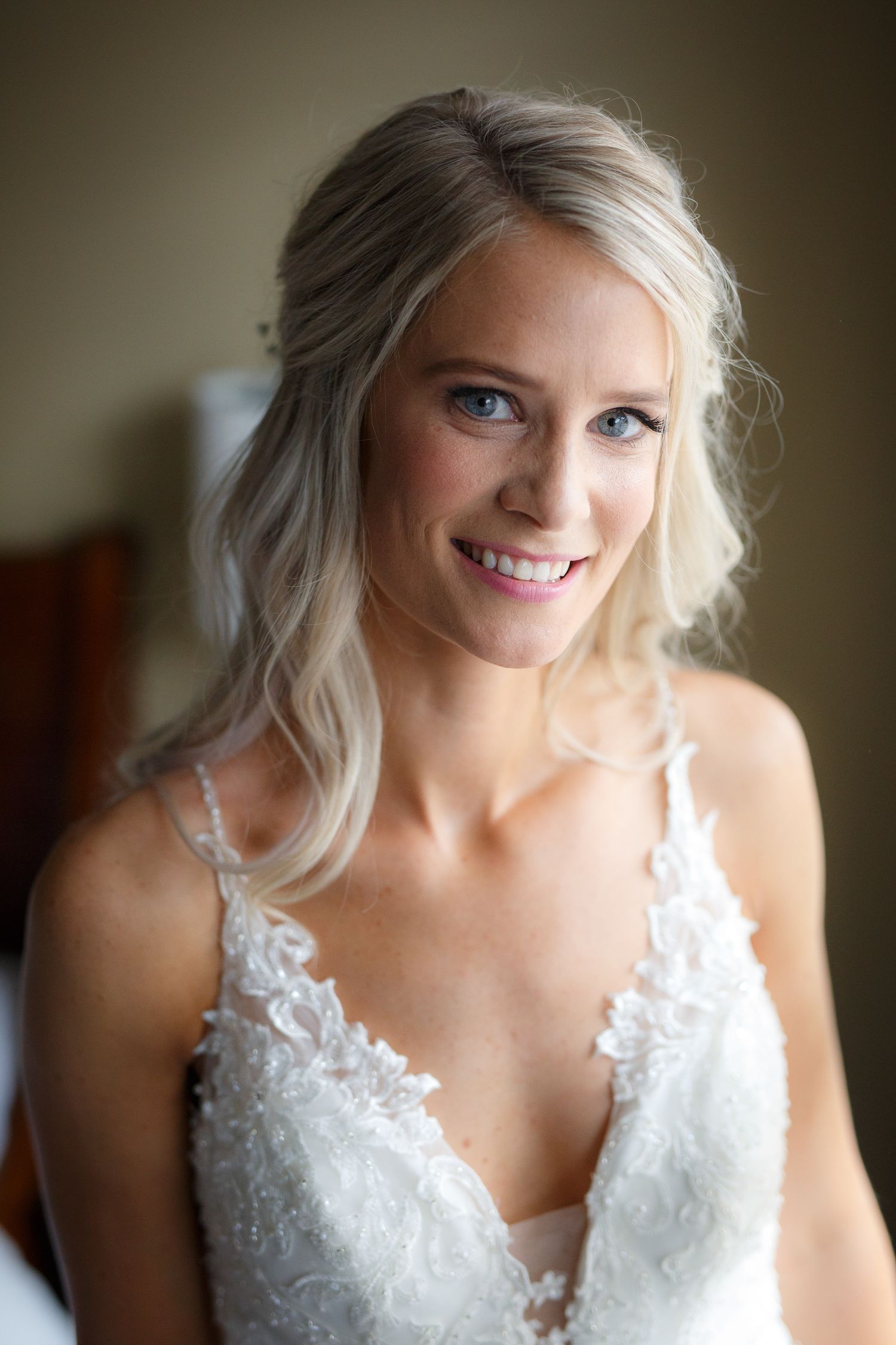 Beautiful bride wearing a white lace wedding dress with delicate straps smiles warmly in soft natural lighting.