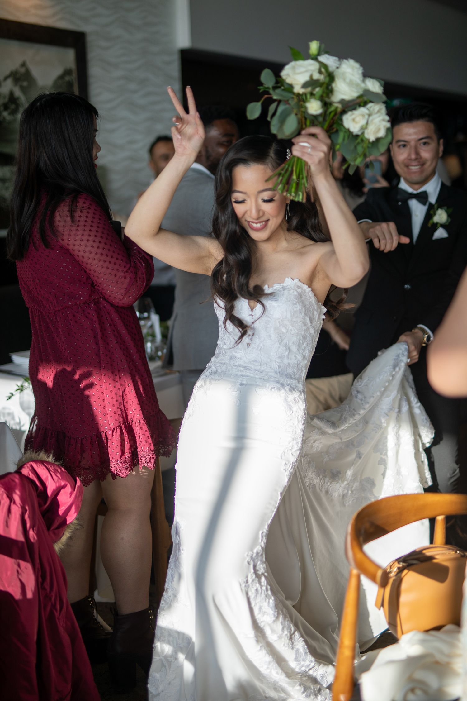 Bride in white strapless gown dancing joyfully holding bouquet of white flowers at wedding reception.