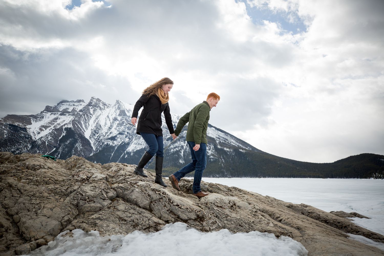 Couple holding hands walking on rocky shoreline with snow-capped mountains and frozen lake in background at Lake Minnewanka in Banff.