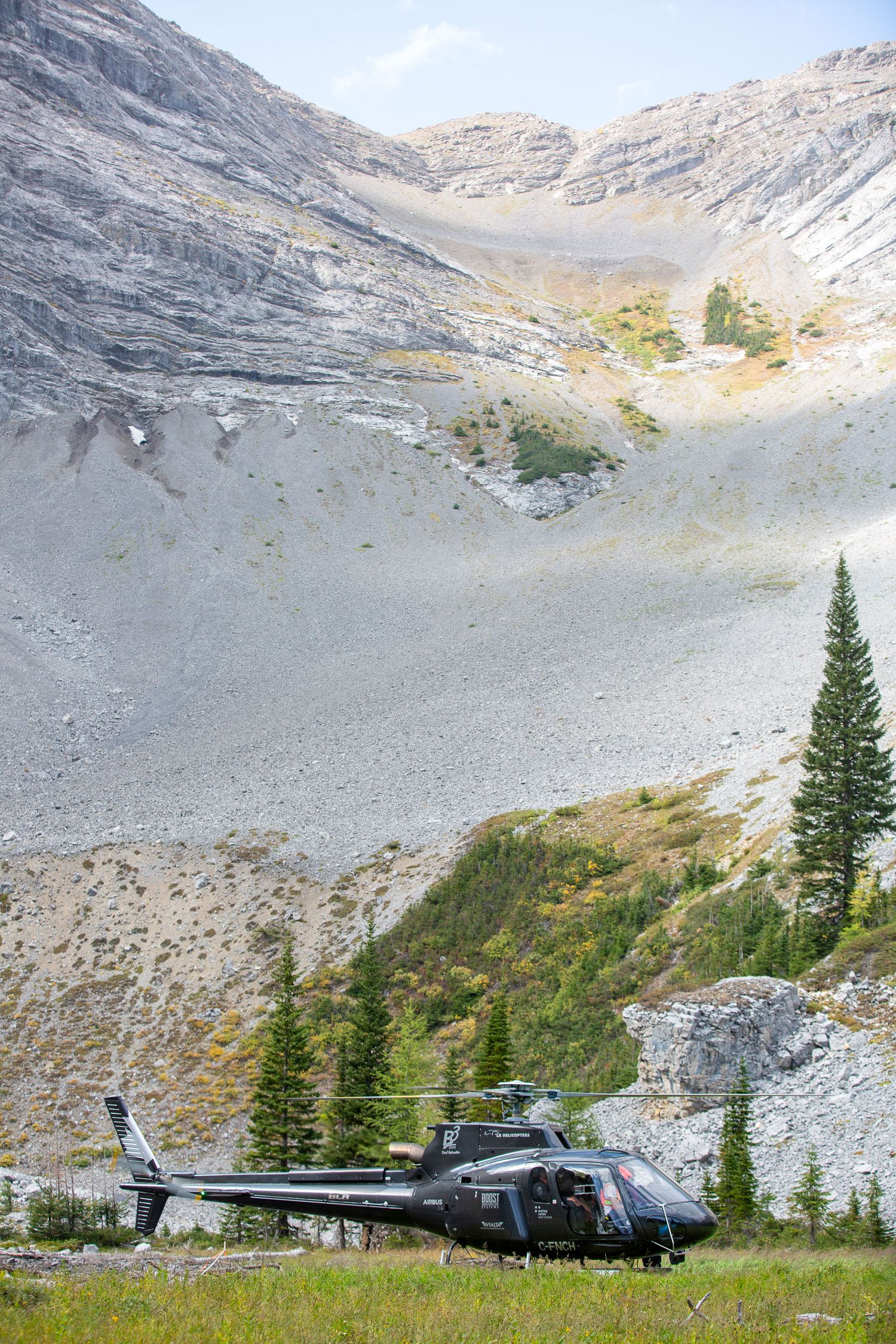 Black helicopter parked in alpine meadow with rocky mountain slopes and evergreen trees in background in Kananaskis, Alberta.