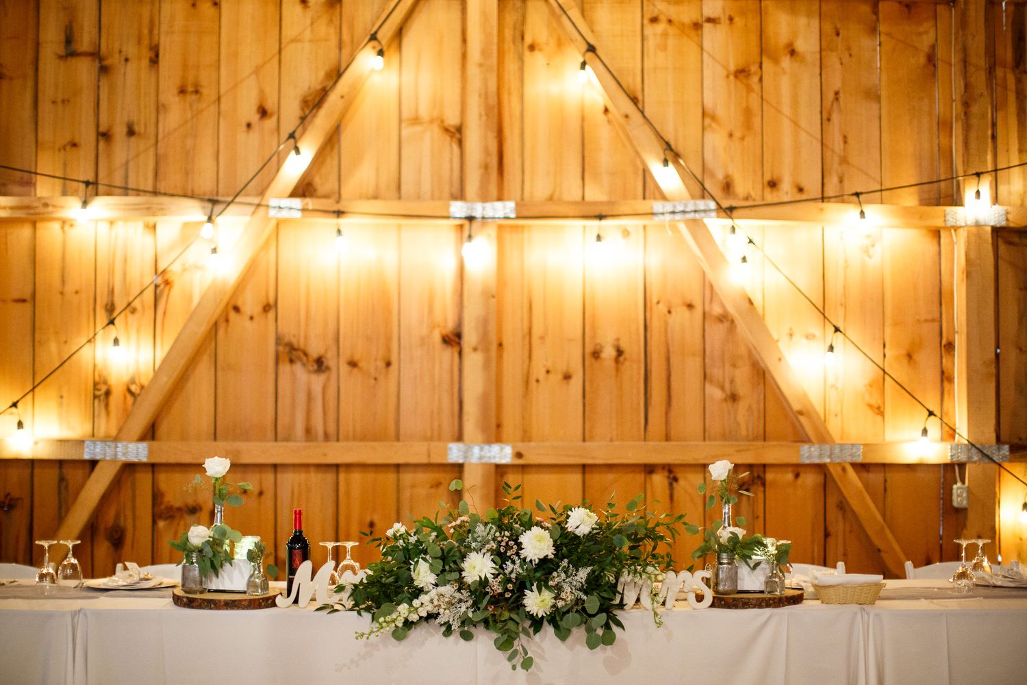 Rustic barn wedding reception with string lights, wooden walls, and elegant floral centerpiece of white roses and greenery Cherry creek