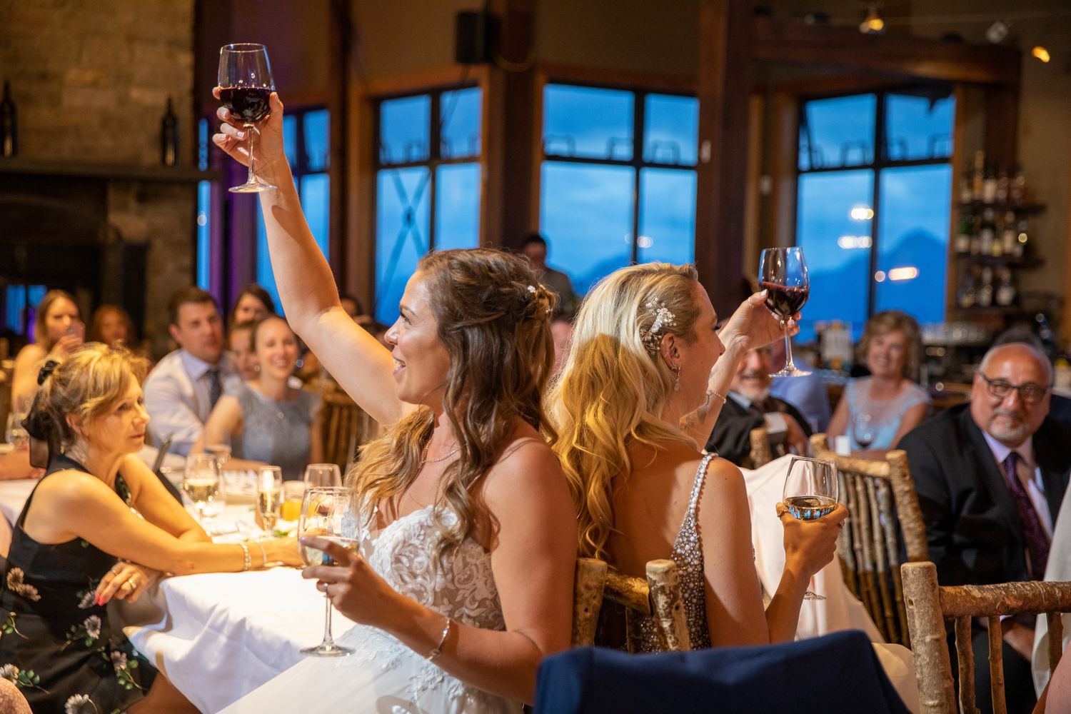 Two brides raise wine glasses in a joyful toast surrounded by wedding guests at an elegant indoor reception at Kicking Horse Resort, BC