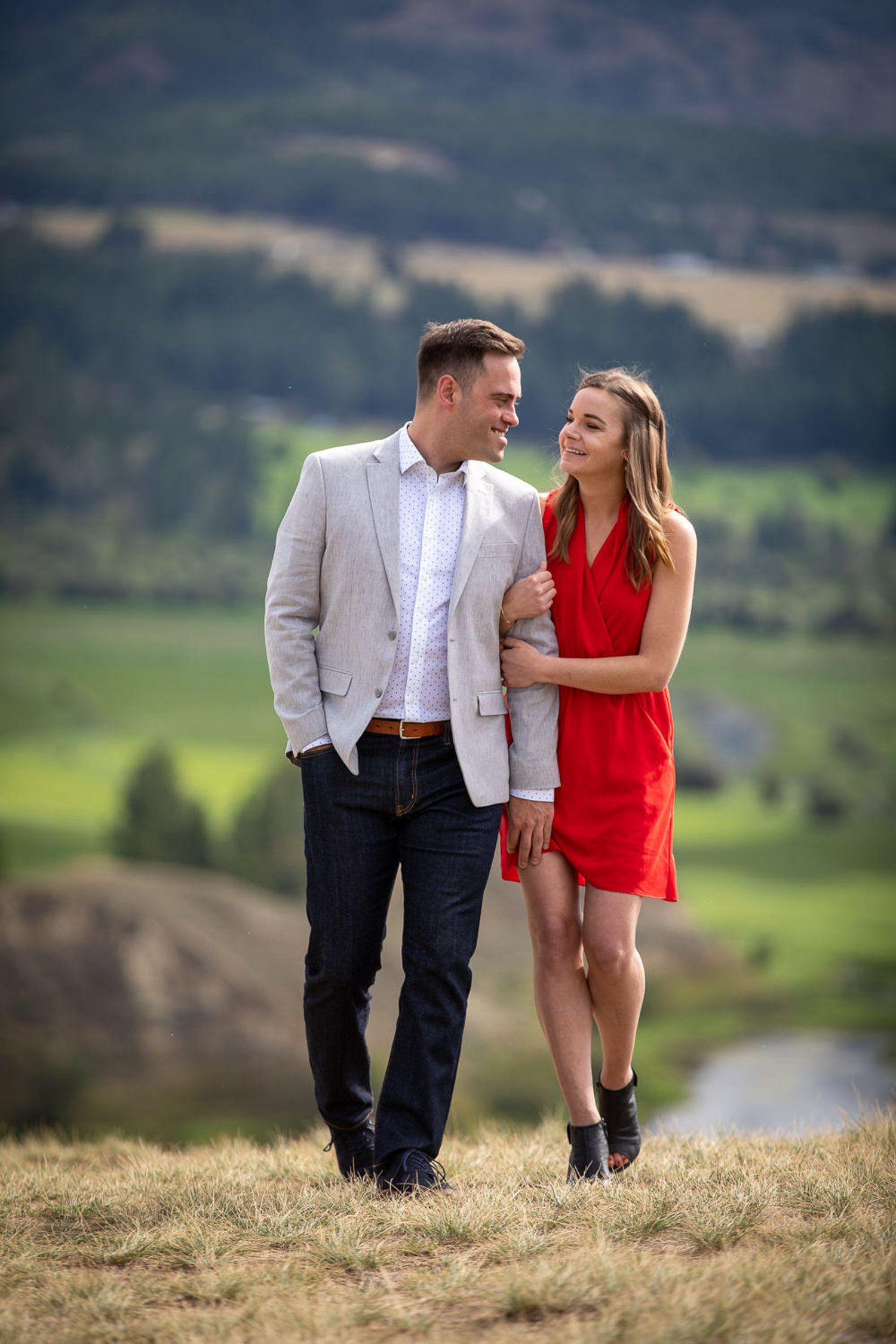 A couple shares a romantic moment on a hilltop overlooking a scenic valley landscape on a sunny day in Wetland Reserve in Invermere, BC.