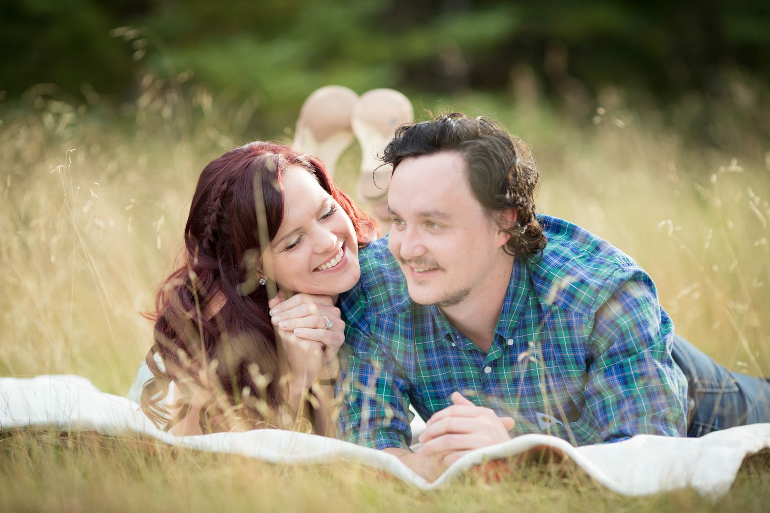 A couple lies on a blanket in tall grass while sharing a romantic moment together in soft natural lighting in Fish Park, Calgary, AB.