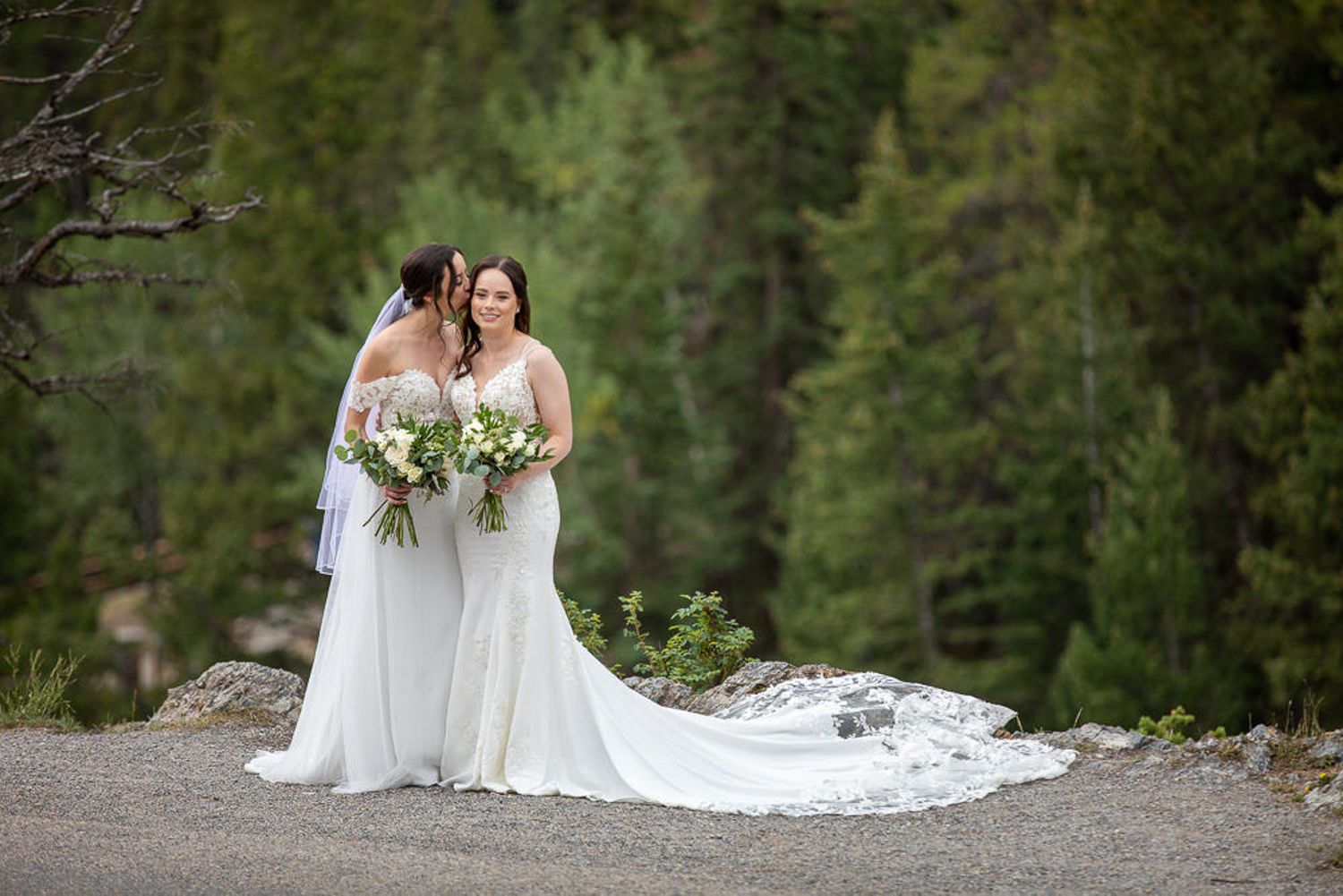 Two brides in elegant white wedding dresses hold bouquets while standing on a mountain path with evergreen forest backdrop in Banff, AB.