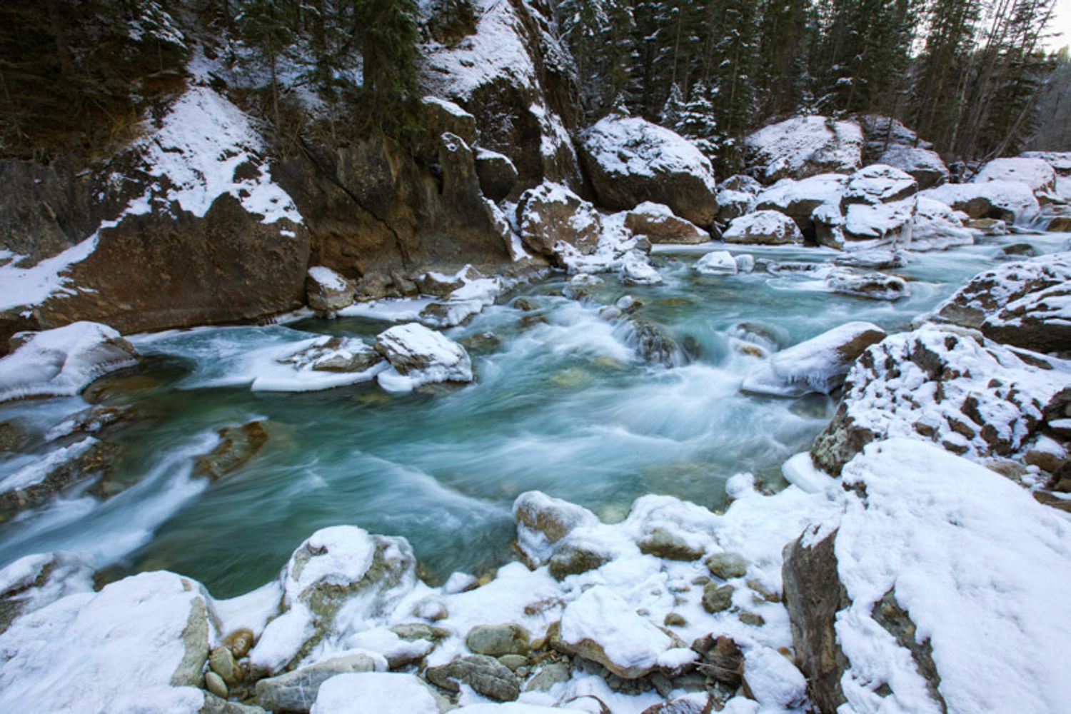 Turquoise river water flows through snow-covered rocks in a winter forest landscape. Toby creek in Panorama, BC.