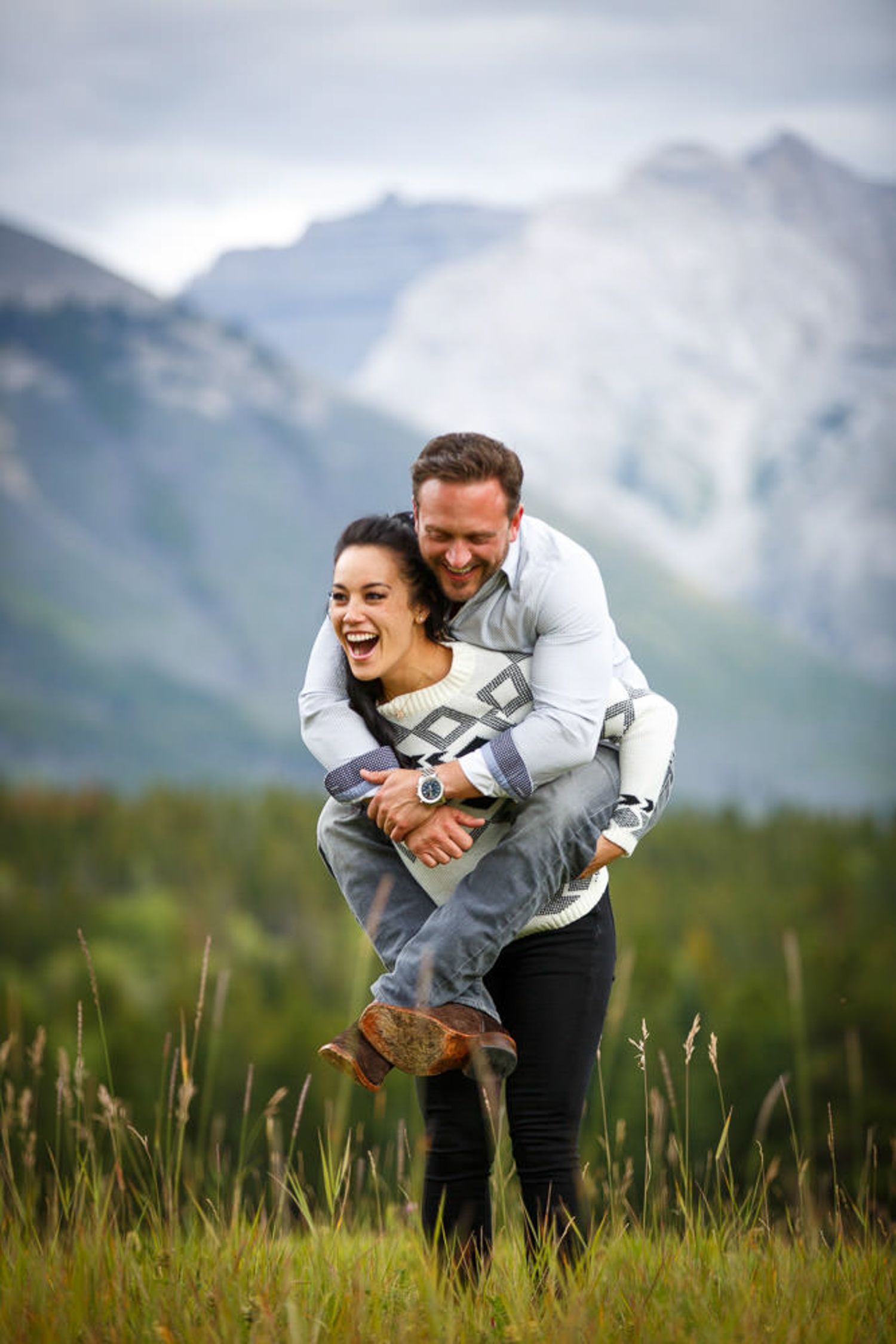 Couple playfully embraces in a meadow with majestic mountain peaks in the background during golden hour in Banff, AB